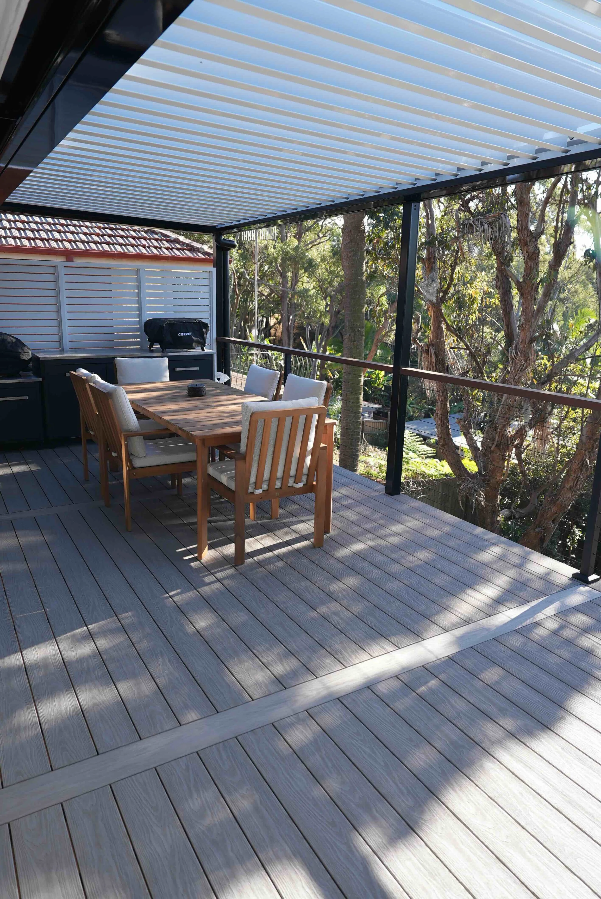 Outdoor patio with wooden dining table and chairs, partially shaded by a slatted metal roof, surrounded by trees and greenery.