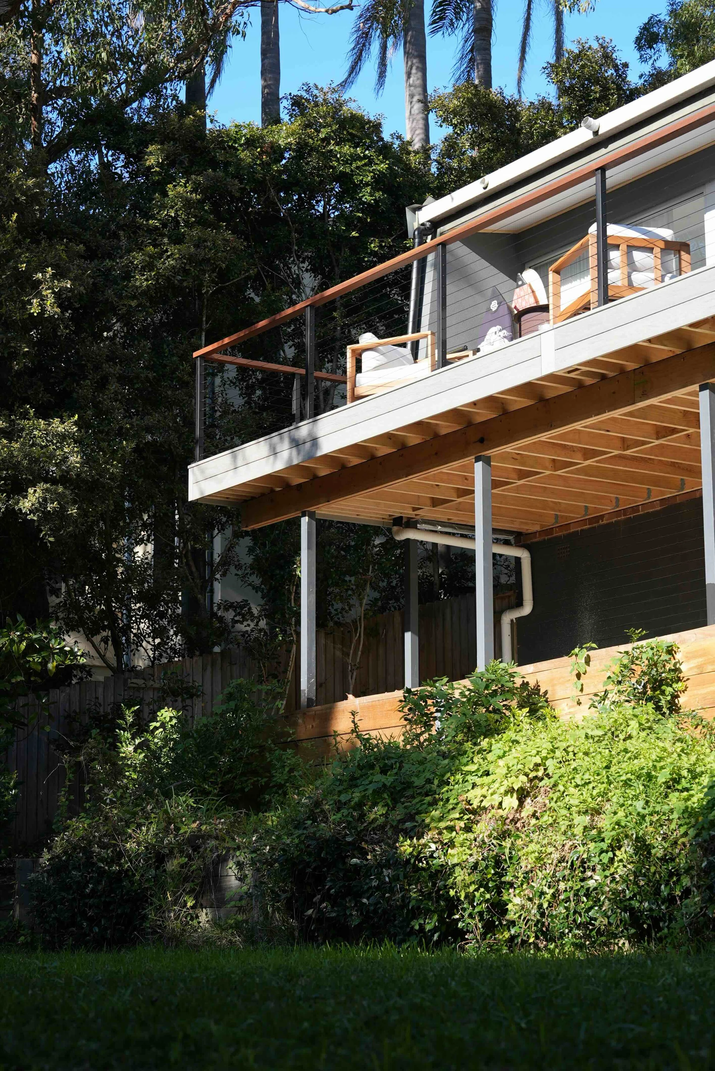 A house with a balcony under construction, surrounded by lush green bushes and trees with a clear blue sky in the background.