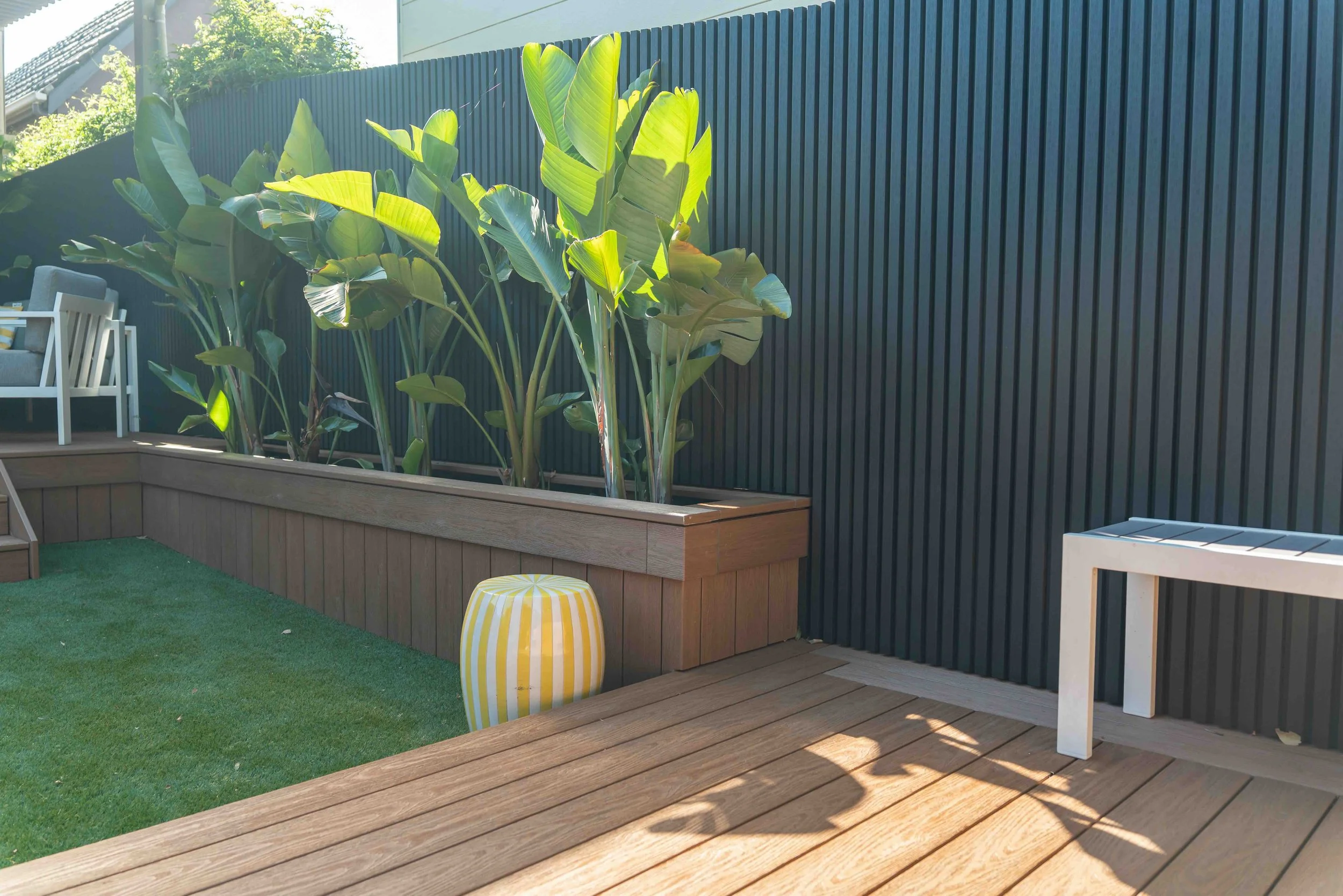 Outdoor patio area with a raised garden bed filled with large green tropical plants, a black vertical slat privacy fence, and a yellow and white striped round stool on a grassy and wooden deck.