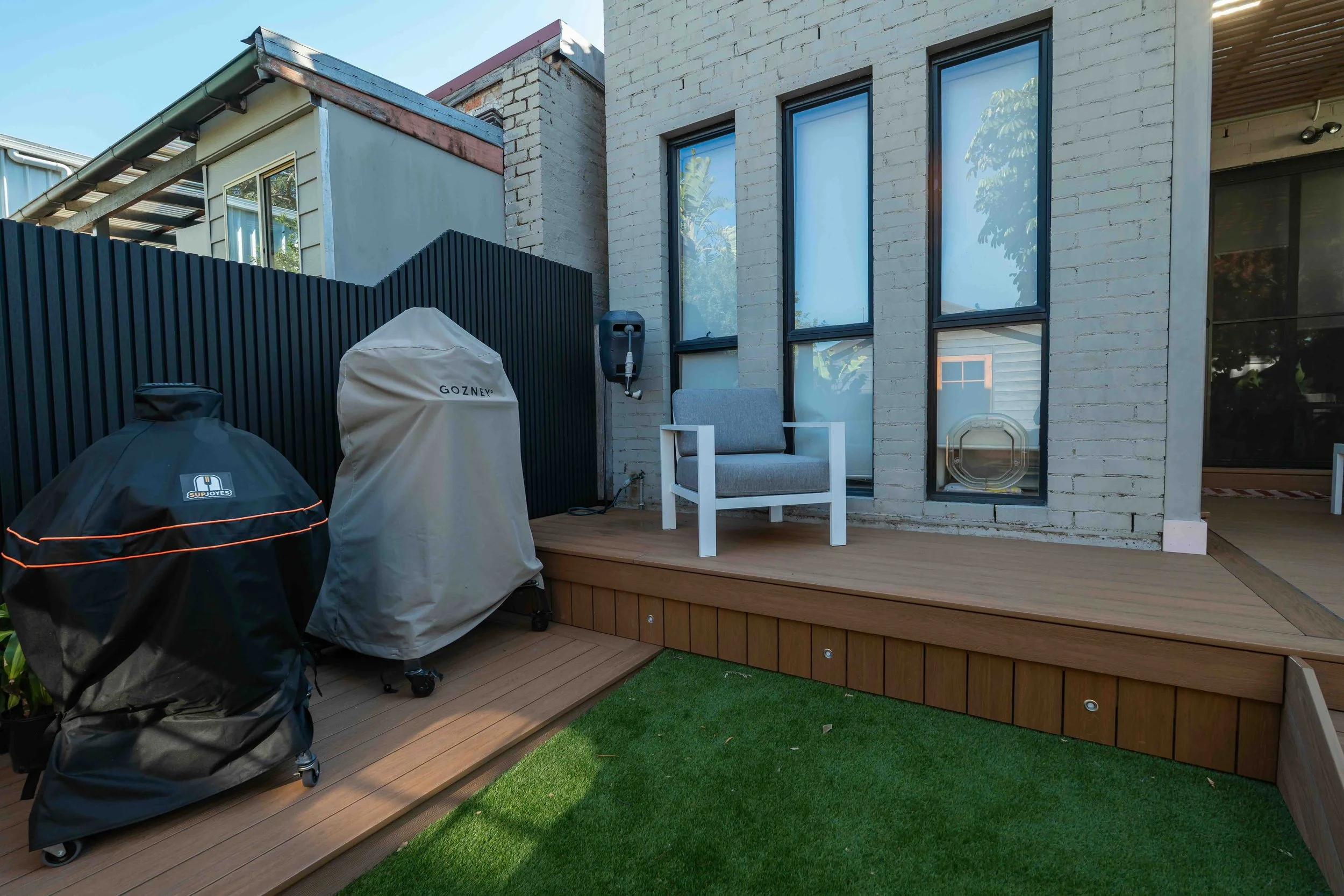 Backyard patio with a wooden deck, a white chair, two covered grills, a black fence, and a house with large windows and beige brick exterior.