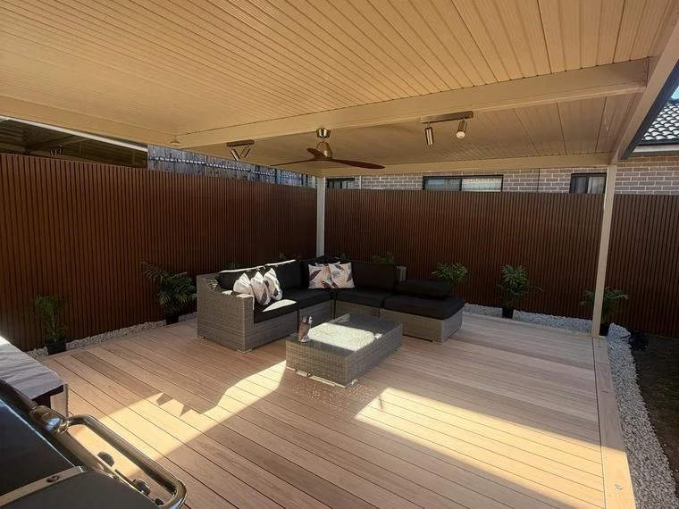 Outdoor patio with a black sectional sofa and throw pillows, a small table, and potted plants, under a covered ceiling with a ceiling fan.