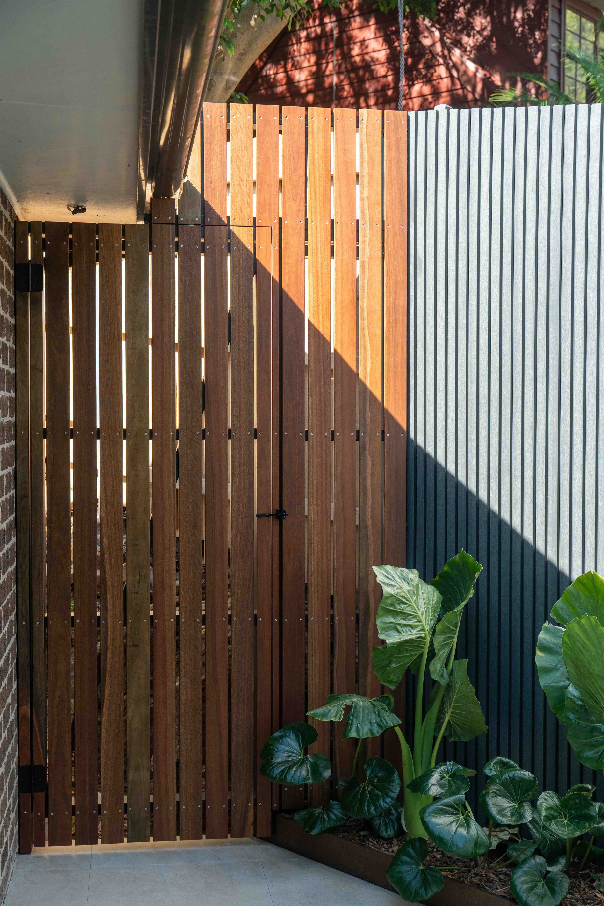 A wooden garden fence with a mix of vertical slats, one side stained darker and the other side lighter, with a small section of metal siding above a brick wall on the left, and a large leafy plant with broad green leaves on the right.