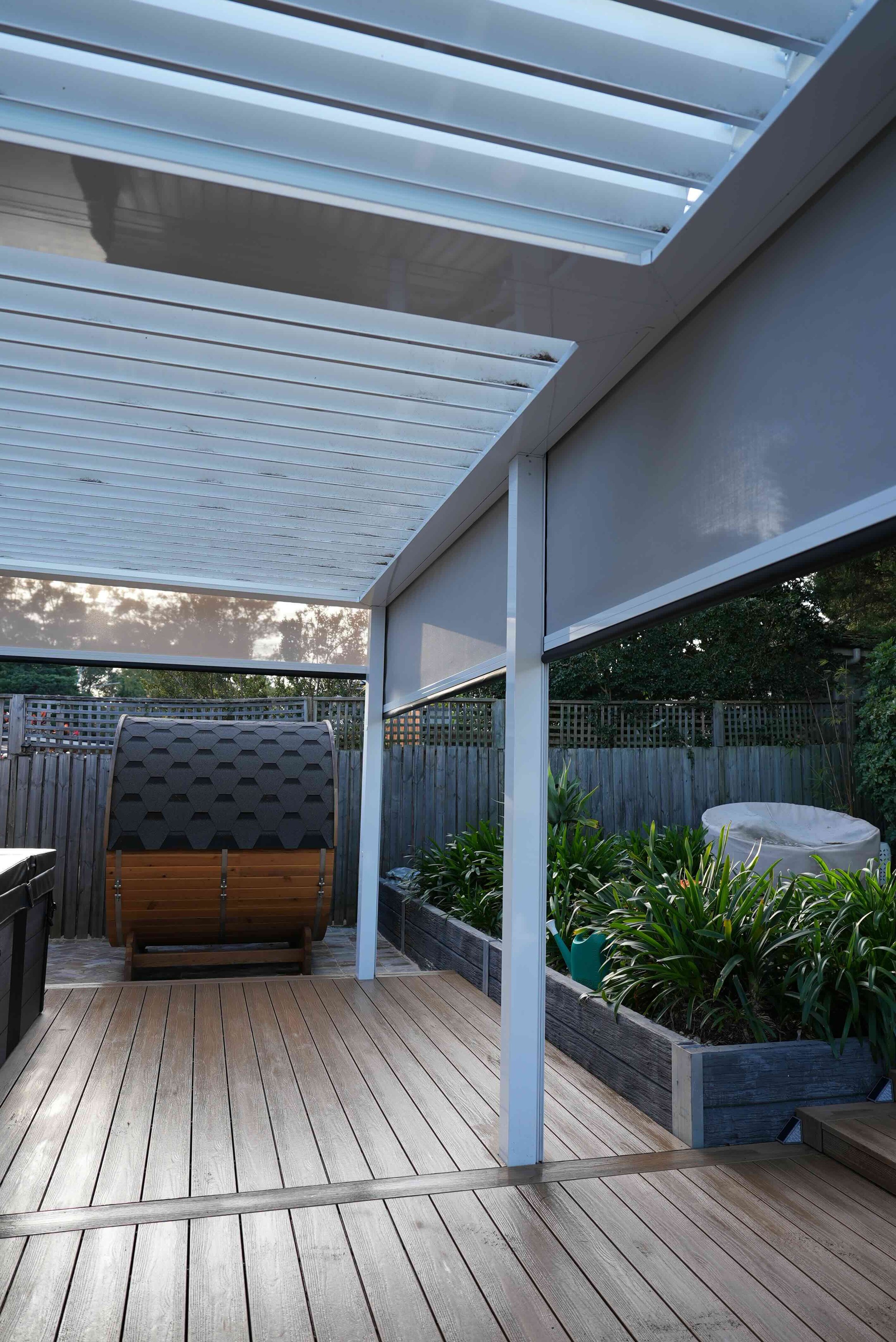 Backyard patio with wood decking, a garden bed with green plants, a wooden privacy fence, and a white pergola and window with blinds, during daytime.