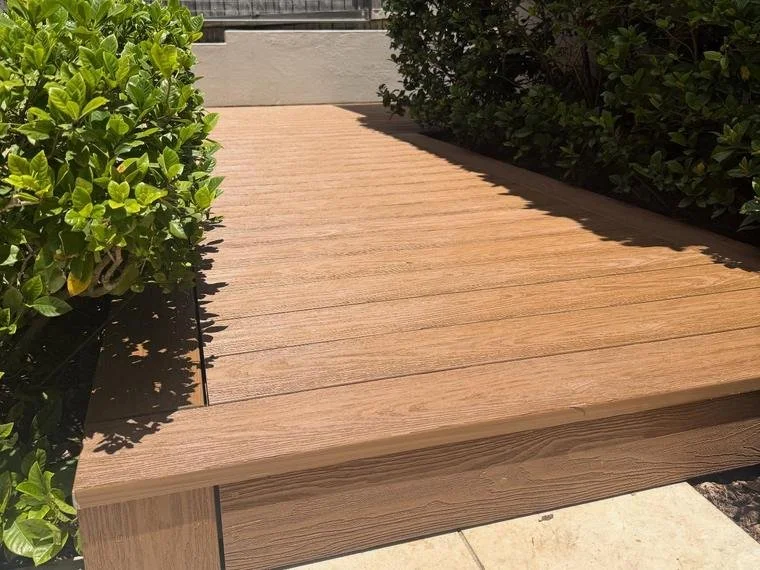 A wooden deck bordered by greenery on both sides, with sunlight casting shadows on the surface.