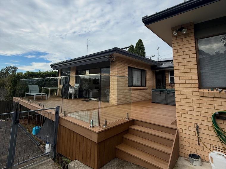 A modern house with a large wooden deck and glass railing, outdoor furniture, and a garden area, under a partly cloudy sky.