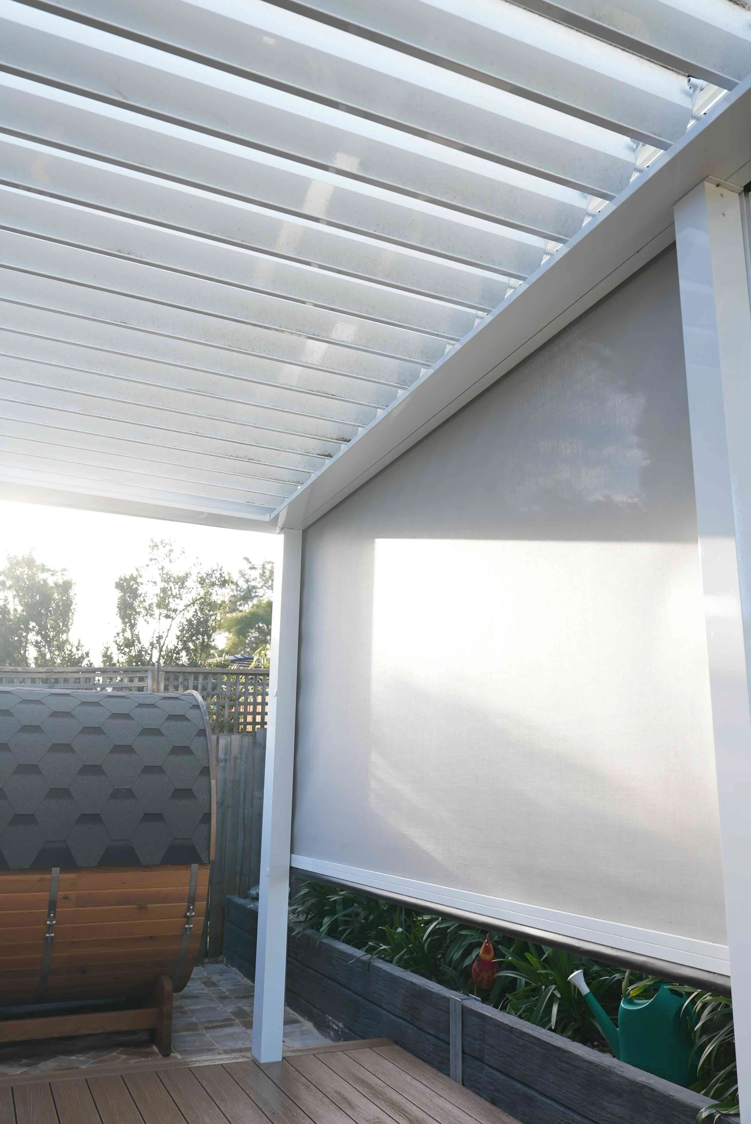 A white outdoor pergola with slatted roof, attached to a house, with a patio and a garden bed with green plants underneath.