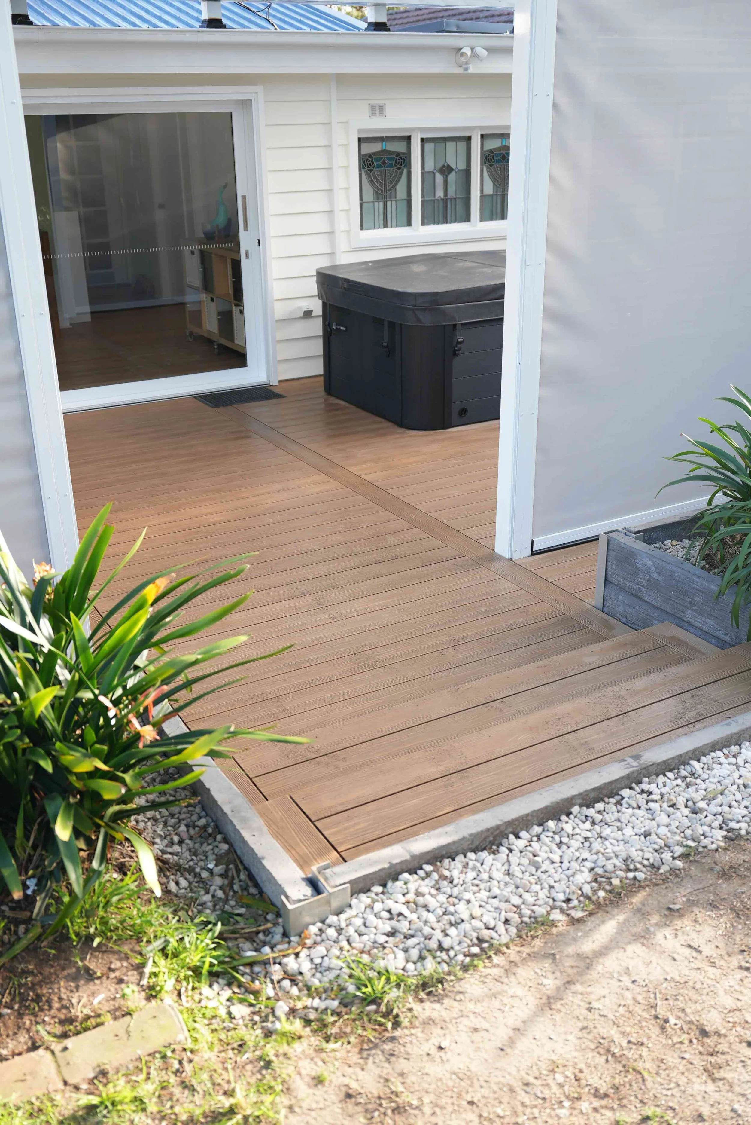 A newly installed wooden deck outside a house with a sliding glass door and a trash bin nearby. The deck is bordered by gravel and some plants, with part of the house's exterior and window visible.