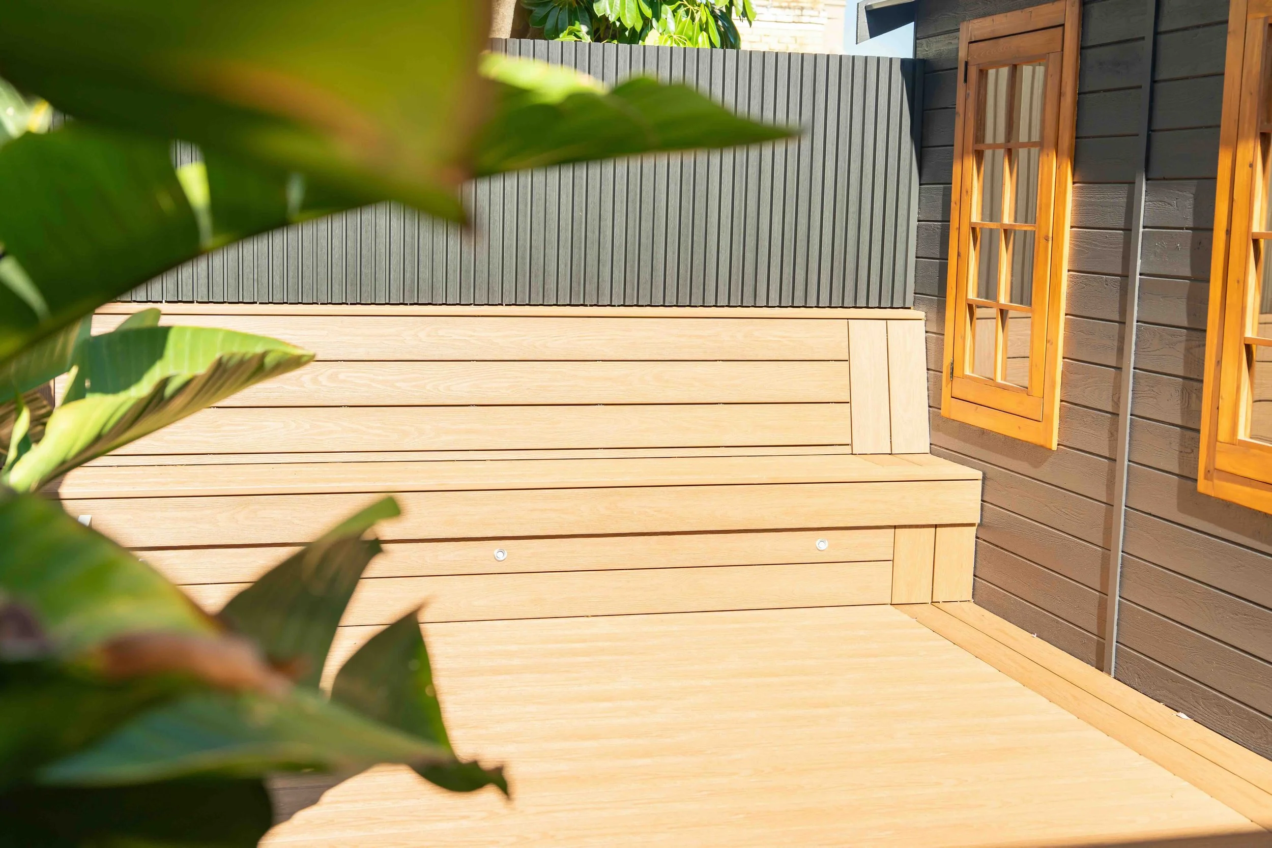 Close-up of green leaves in foreground with a modern wooden deck and gray and brown exterior walls of a building in the background.