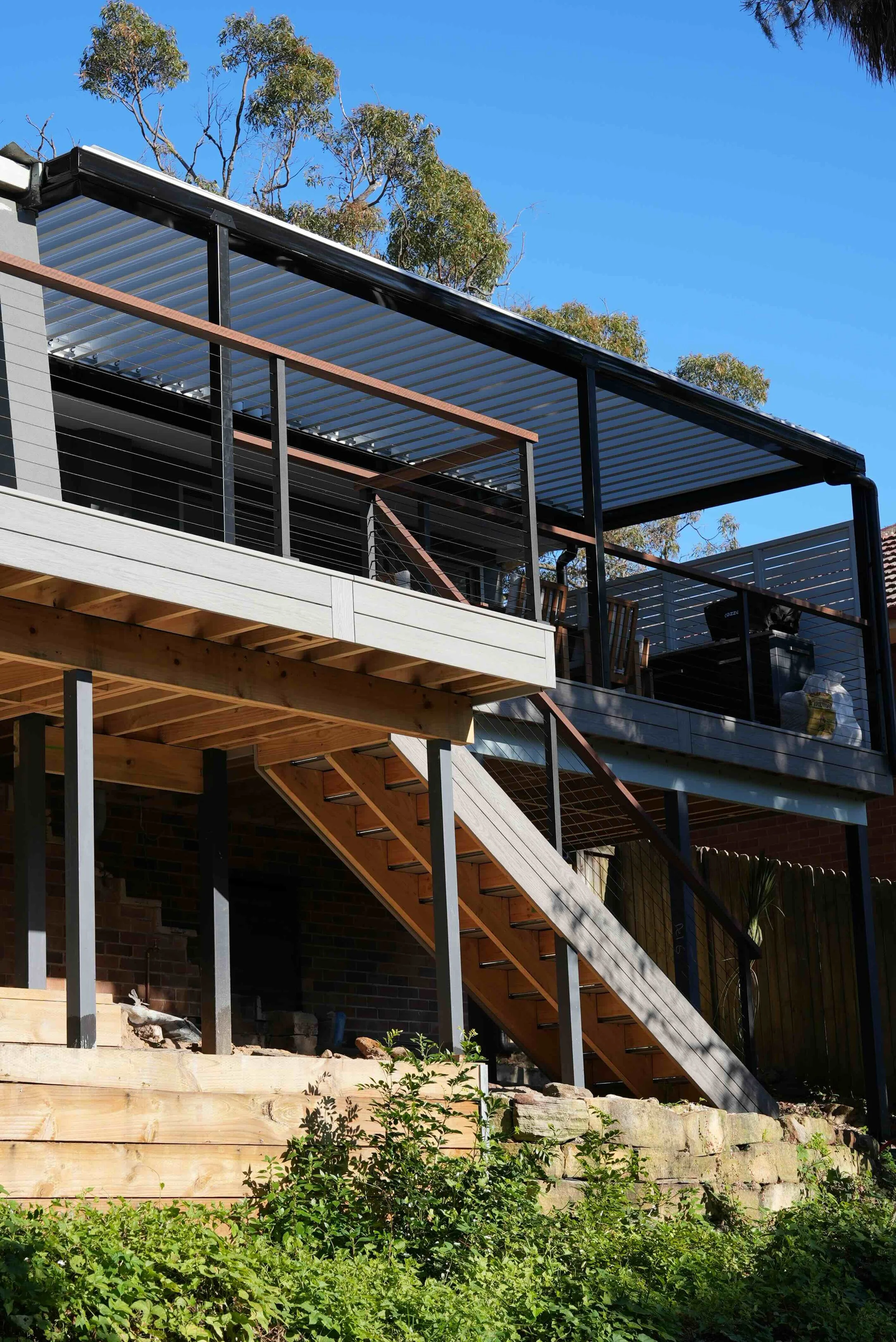 Construction site of a two-story building with a balcony, wooden stairs, and metal railings, under a clear blue sky with trees in the background.