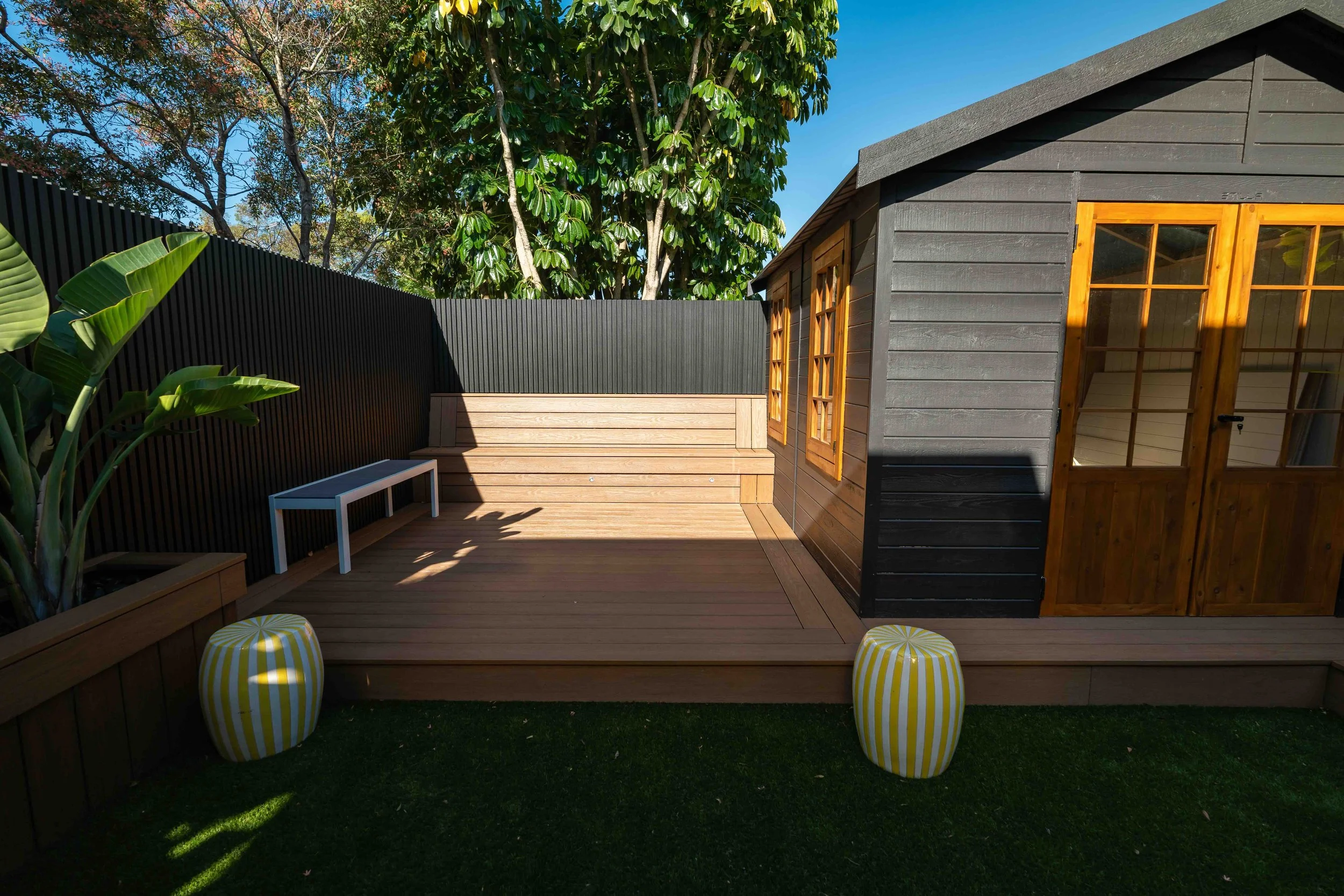A backyard patio with wooden decking, two yellow and white striped poufs, a black privacy fence, a gray wooden shed with windows, and lush green trees and plants under a clear blue sky.