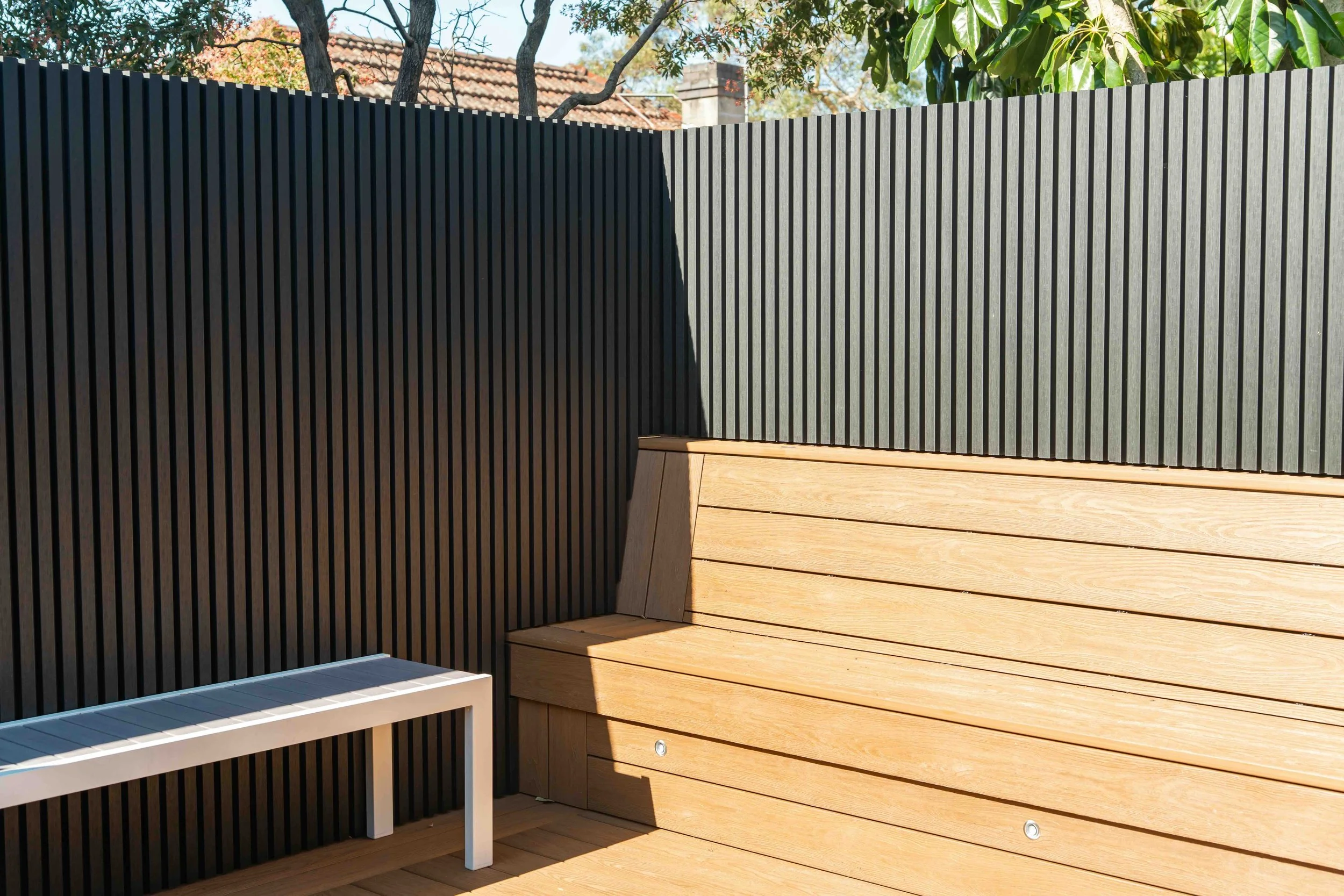 View of a backyard fence with black vertical slats, a wood bench, and a wooden wall, with trees and a house roof in the background.