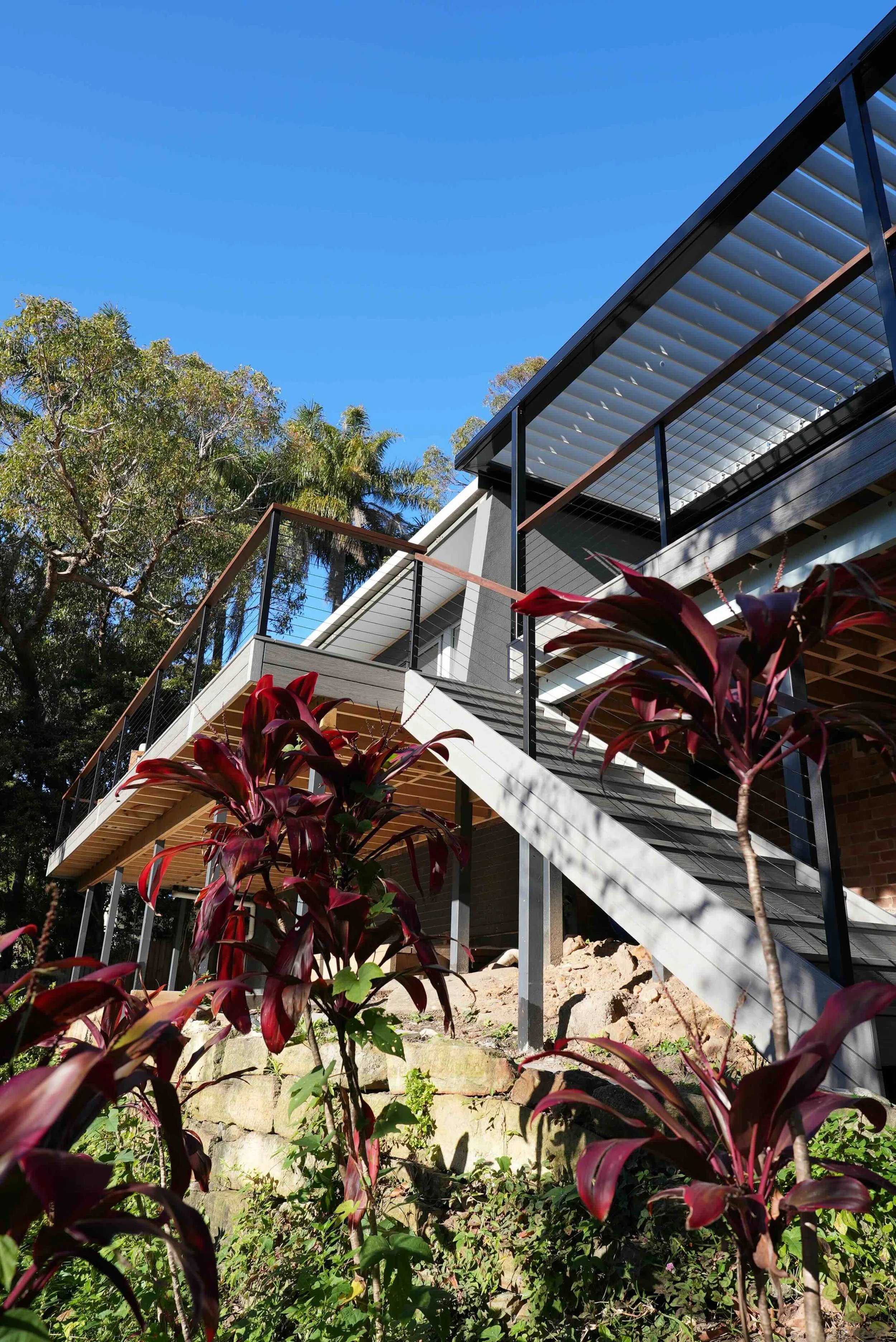 Modern house with large balcony and metal railing, surrounded by green trees and plants, under a clear blue sky.