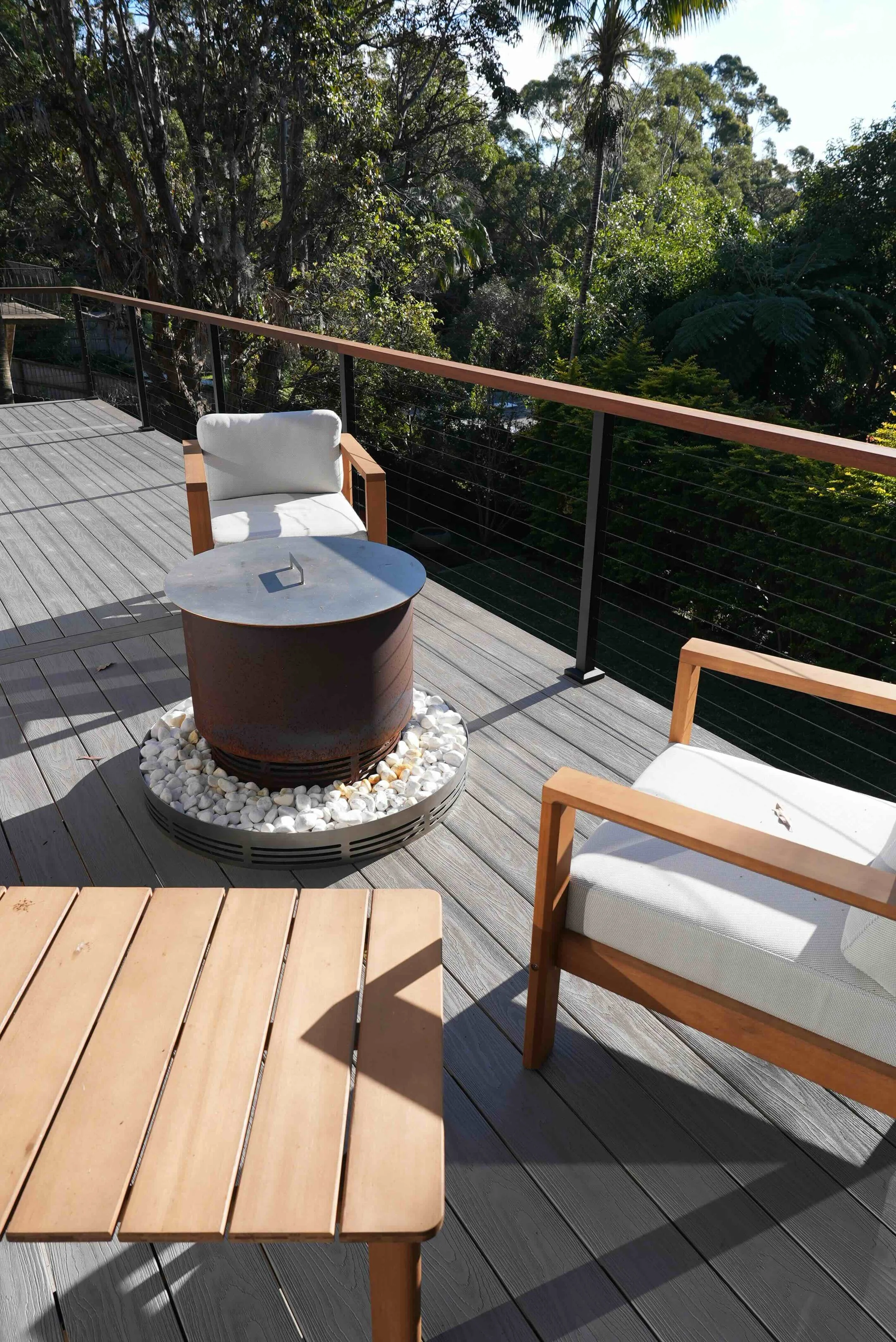 Balcony with wooden chairs and a round fire pit filled with white pebbles, overlooks lush green trees under clear daylight.