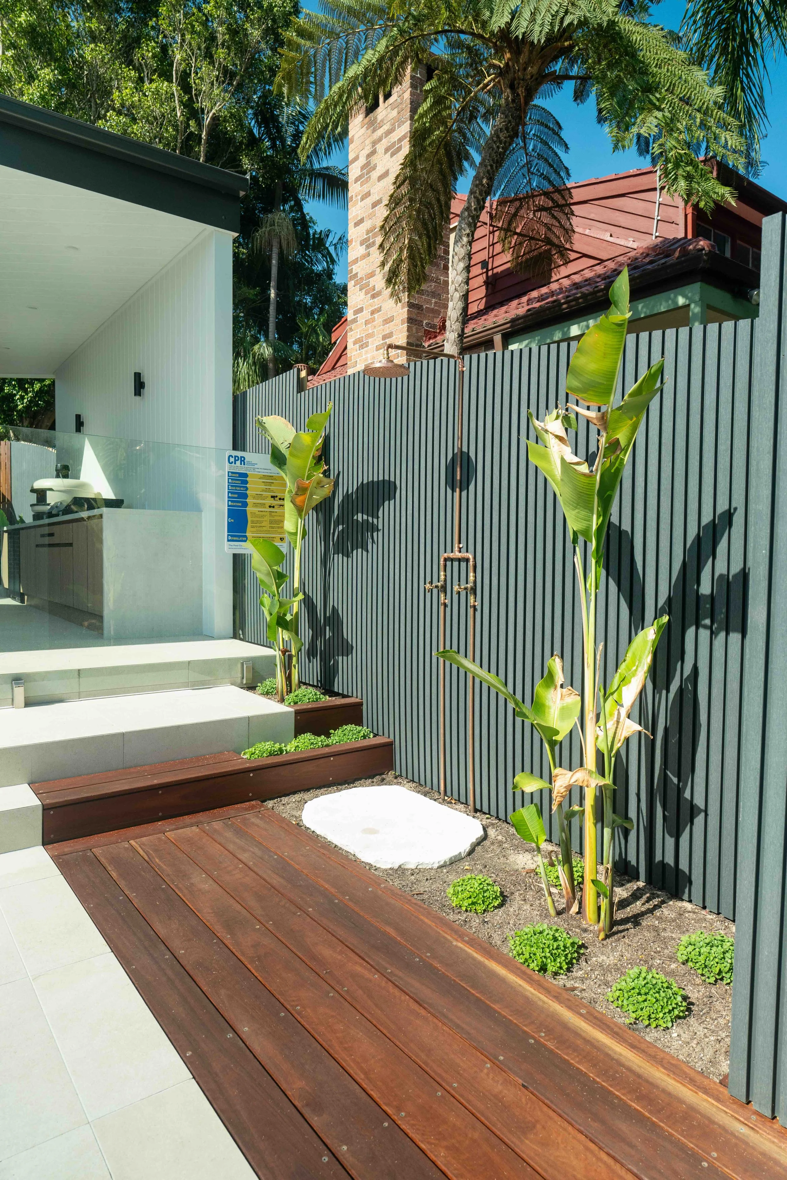 Modern outdoor backyard area featuring a black vertical slat fence, small trees, and tropical plants including banana trees. There is a concrete pathway, a wooden deck, a step, and a section of polished concrete or tile. In the background, a house with a brick chimney, a red roof, and an overhang. The sky is clear blue with trees behind.