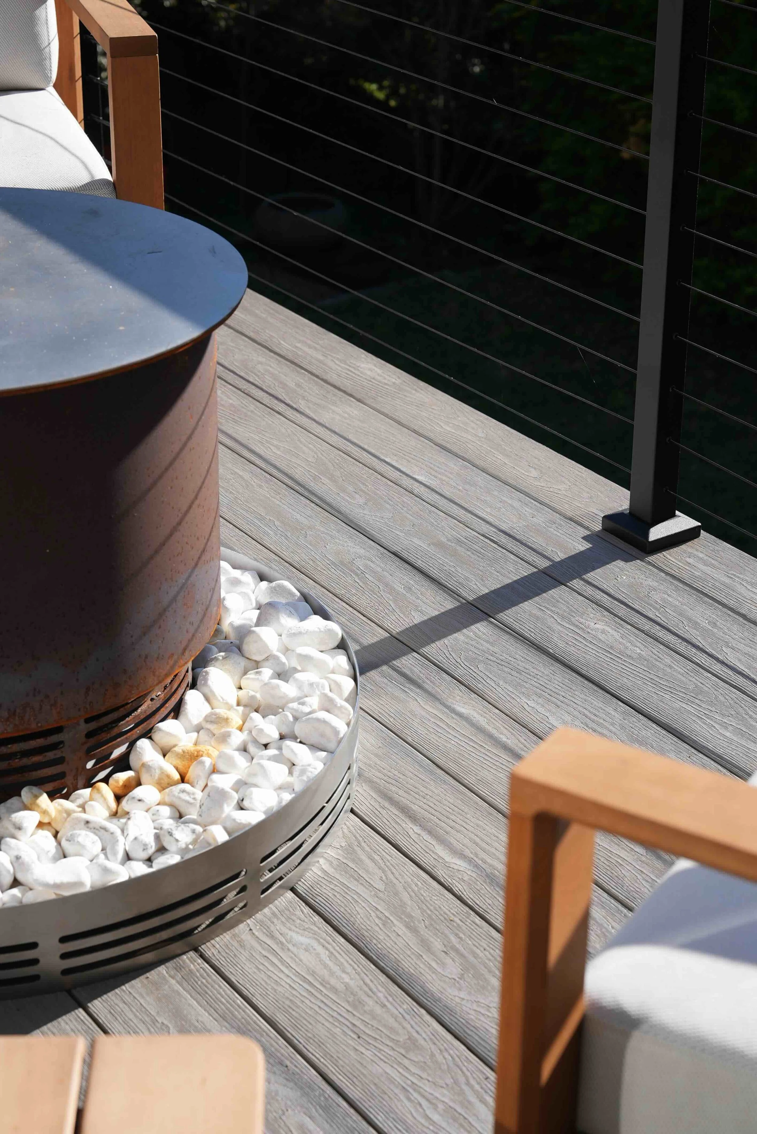 Part of an outdoor patio or balcony with a round table, white stones in a round container, wooden chairs, and a black metal railing.