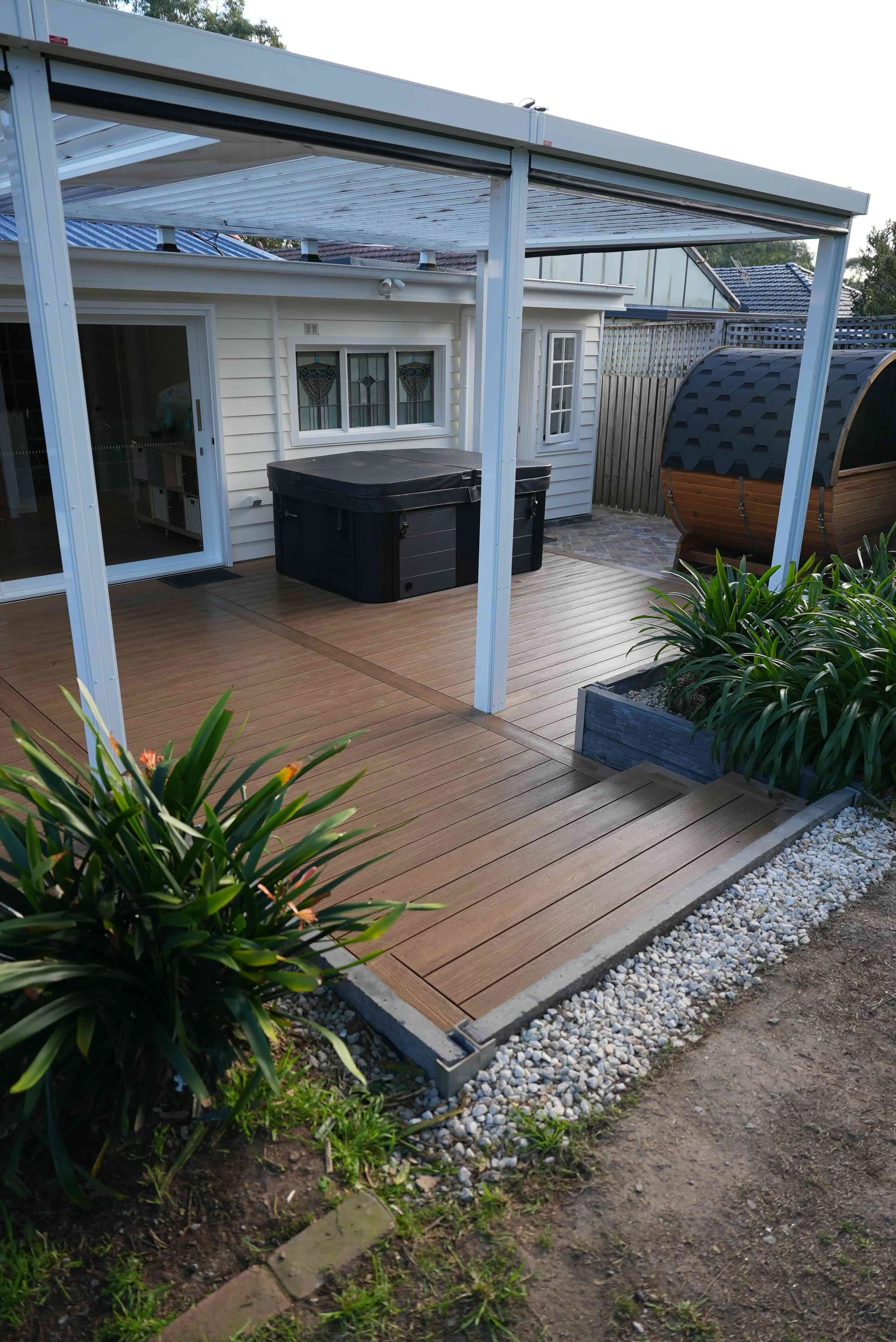 A backyard patio with a wooden deck, a hot tub, and a shaded area with a metal frame structure. There are plants in the foreground and a fence in the background.