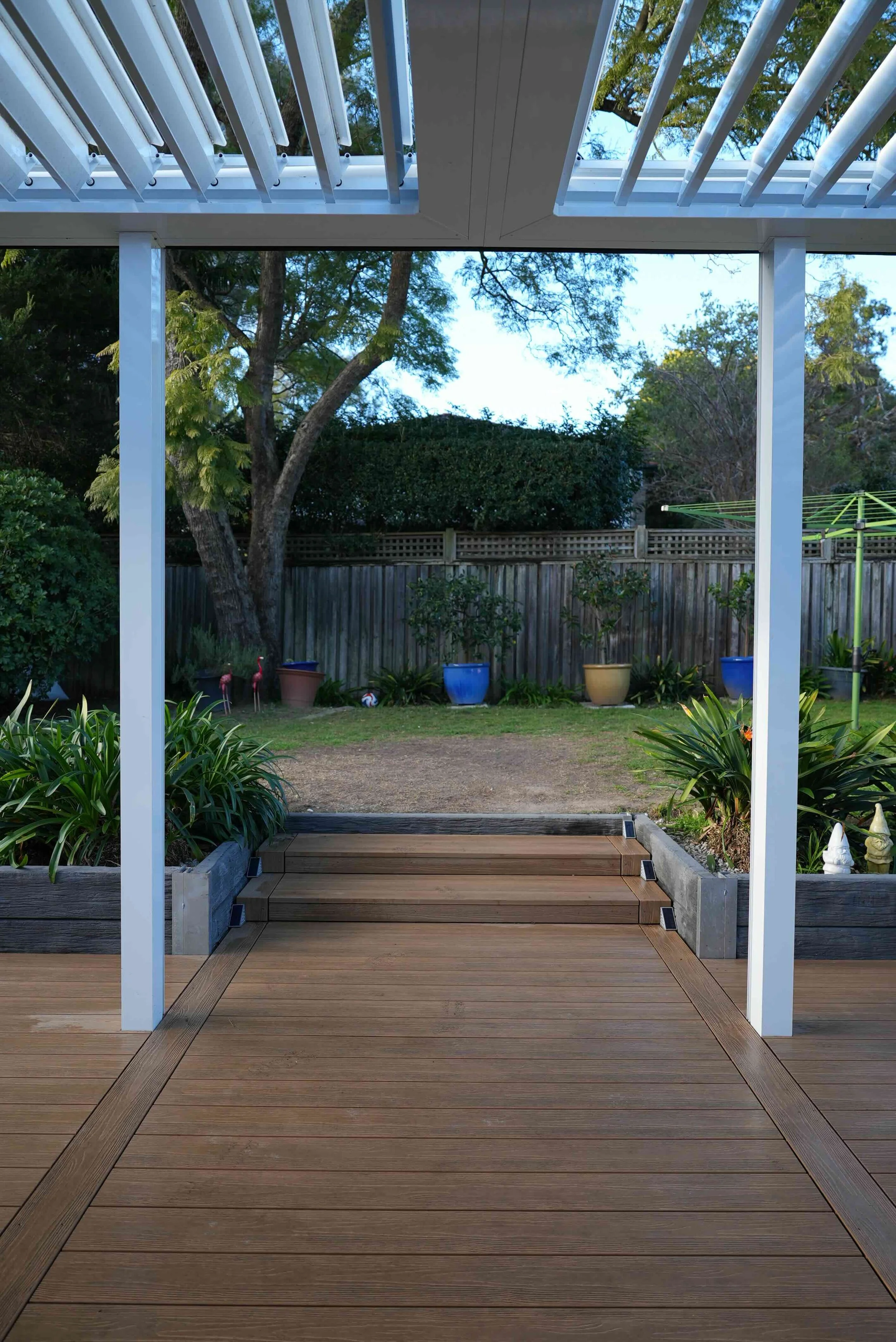 A backyard garden viewed from a porch with wooden stairs and white columns. The garden has potted plants, a lawn, trees, and a wooden fence in the background.
