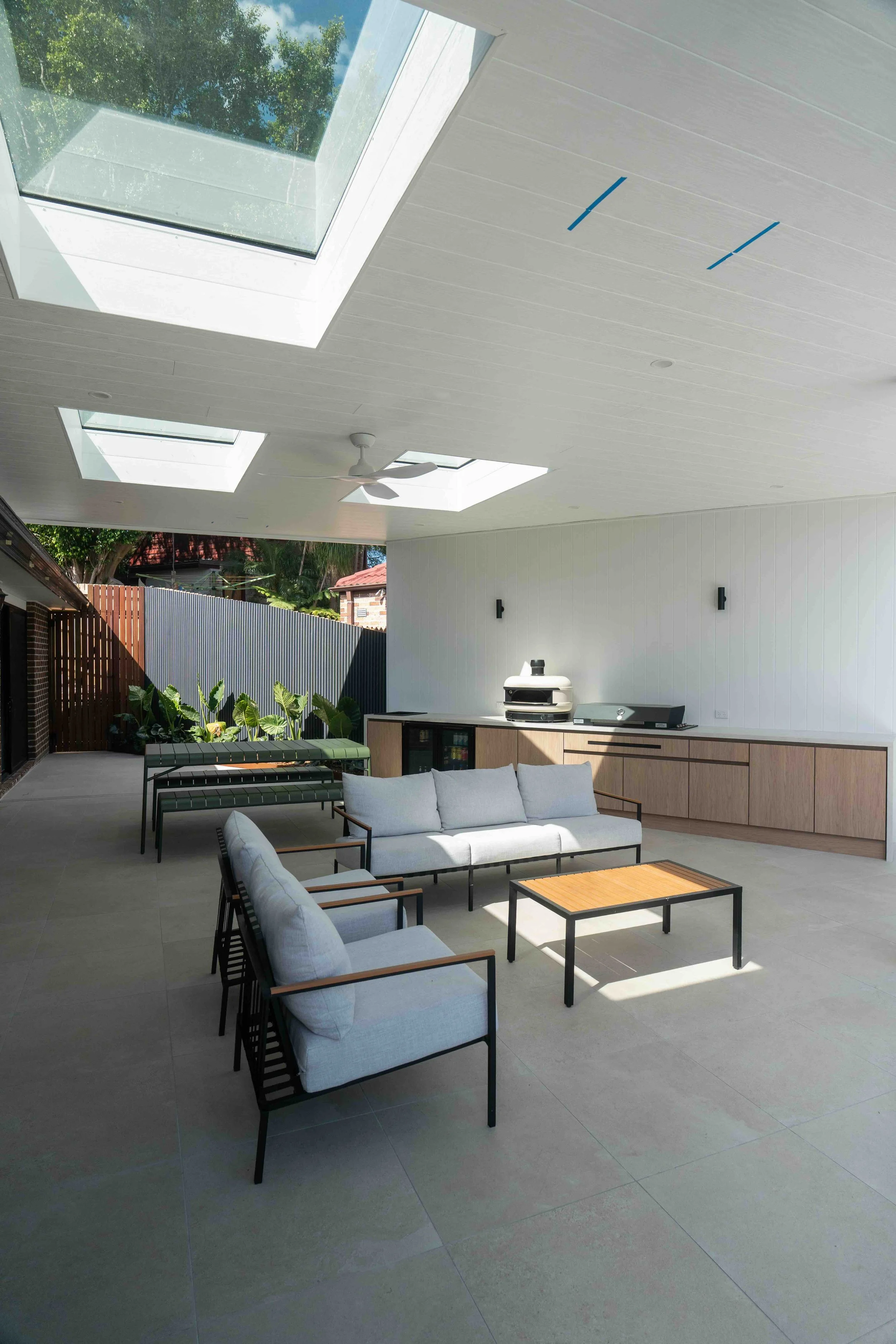 Outdoor patio with modern furniture under a white ceiling that has three skylights and a ceiling fan. There is a gray tile floor, a white wall with black lights, an outdoor grill, a wooden countertop, and a fence with greenery and a house in the background.