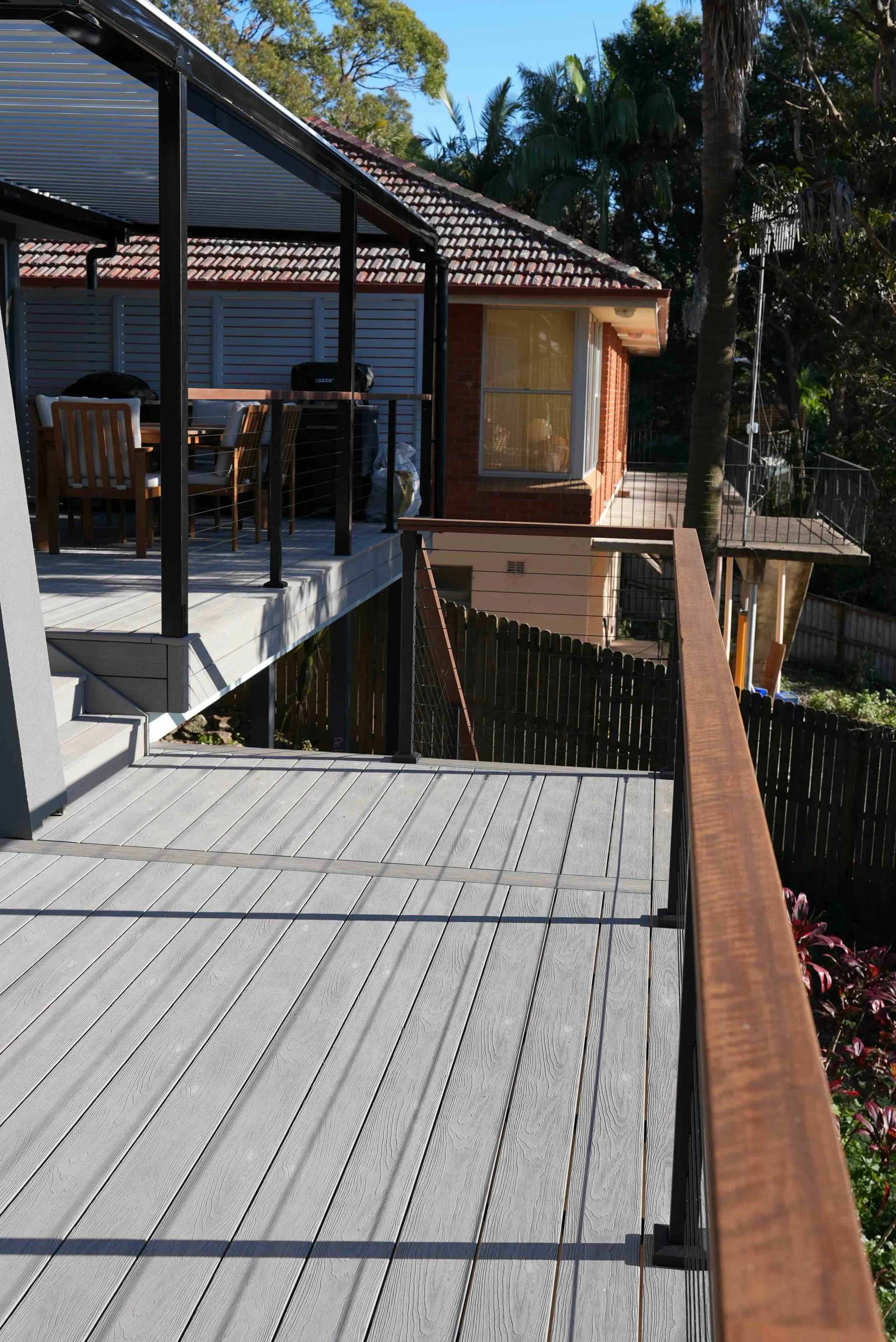 A residential backyard deck with a gray wooden floor and brown railing, overlooking a backyard with trees, a fence, and part of a house with brick walls and large windows.