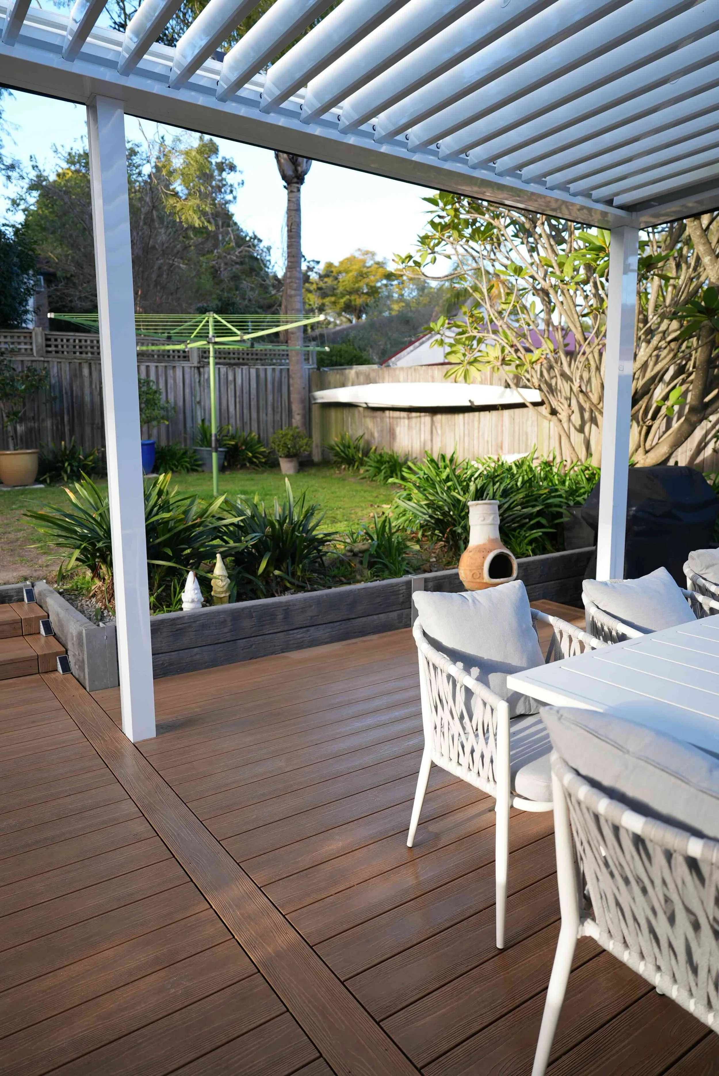 View of a backyard patio with brown wooden decking, white outdoor furniture, potted plants, a fence, trees, and a round above-ground pool in the background.
