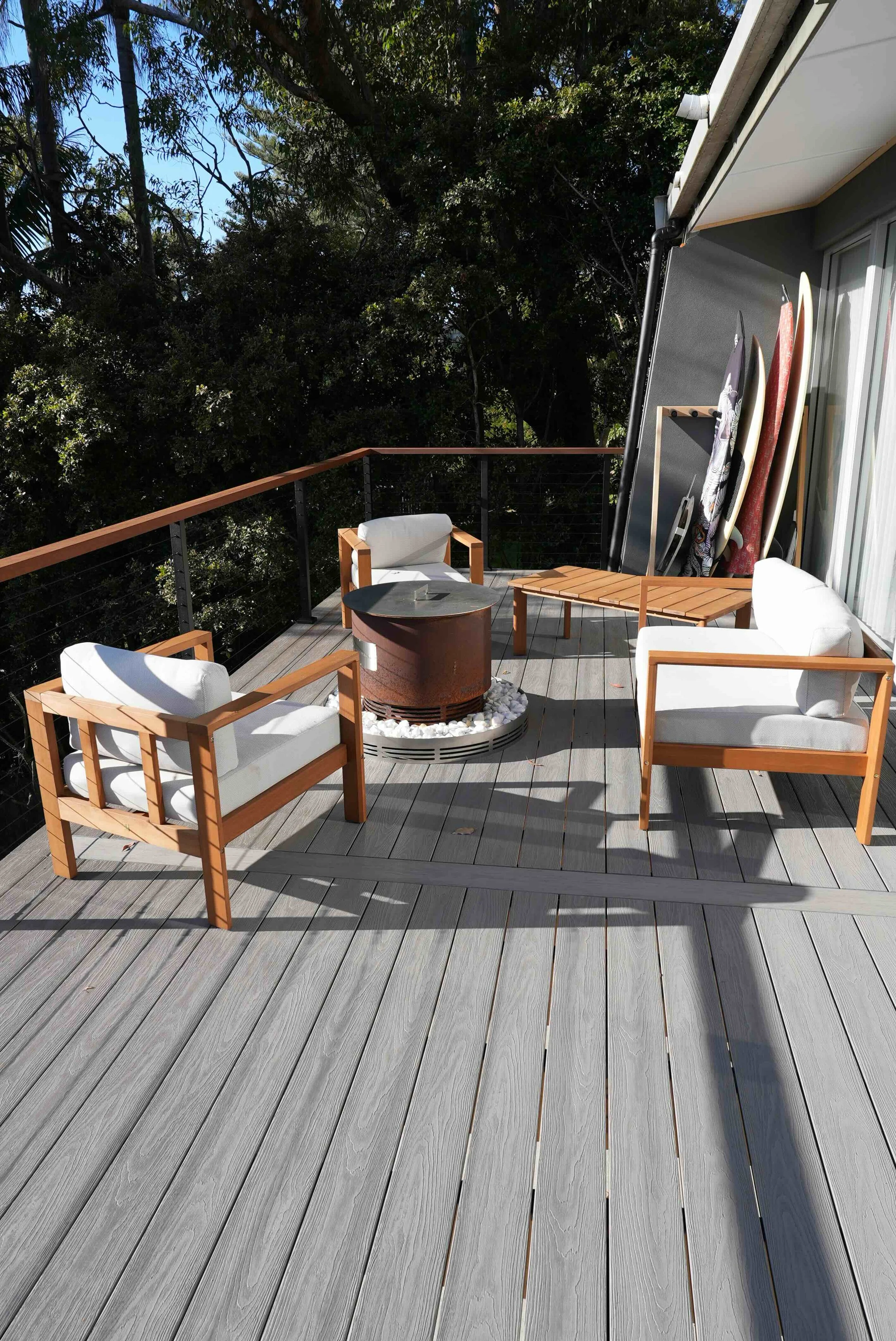 An outdoor patio with wooden furniture, including chairs with white cushions, a small table, and two surfboards standing against the house wall. The patio has gray wooden flooring and is surrounded by lush green trees.