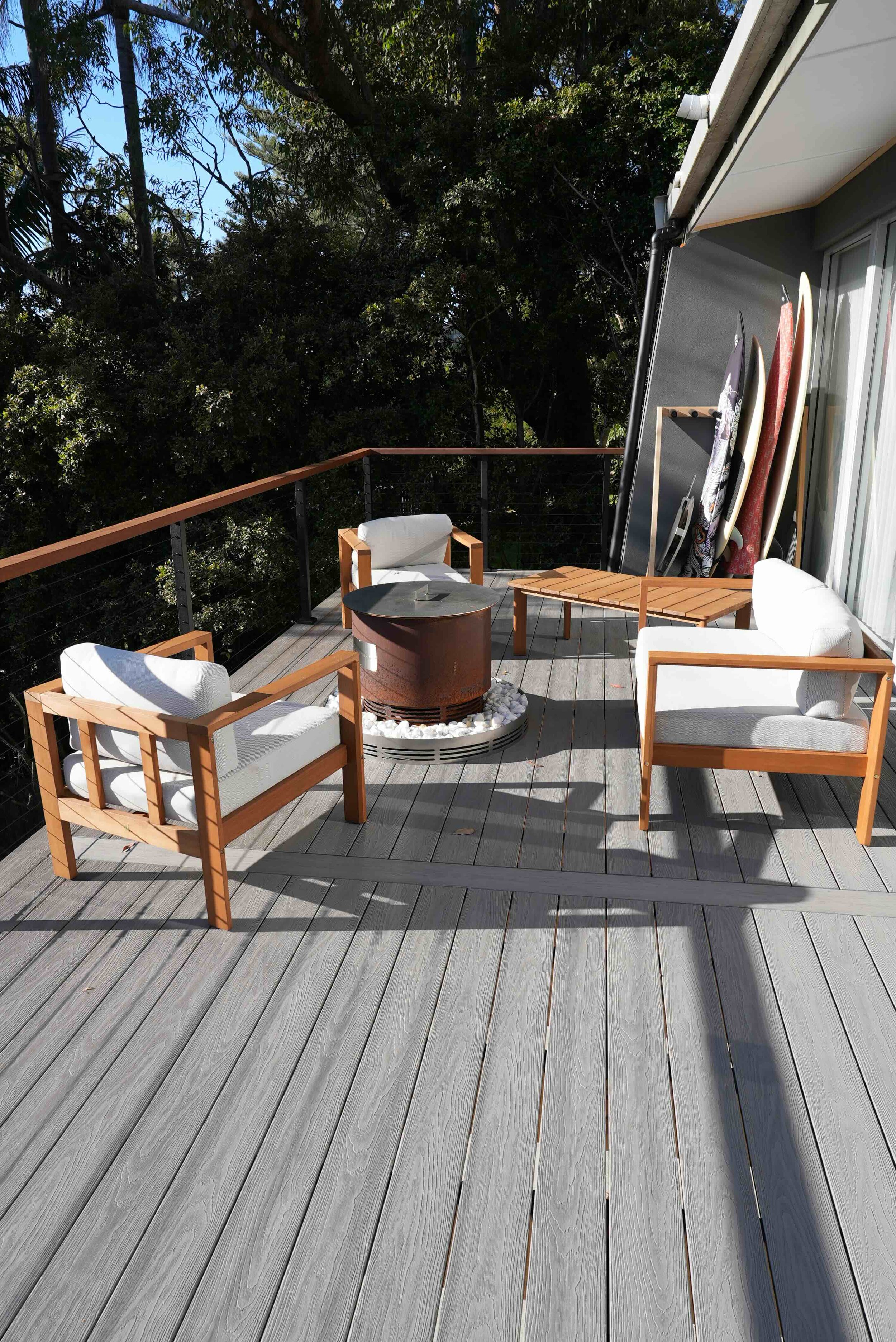 A balcony with modern outdoor furniture, including a round fire pit, four white cushioned chairs with wooden frames, a wooden side table, and surfboards stored upright against a dark exterior wall. The balcony has gray wooden flooring, and a lush green treetop backdrop with a clear blue sky.