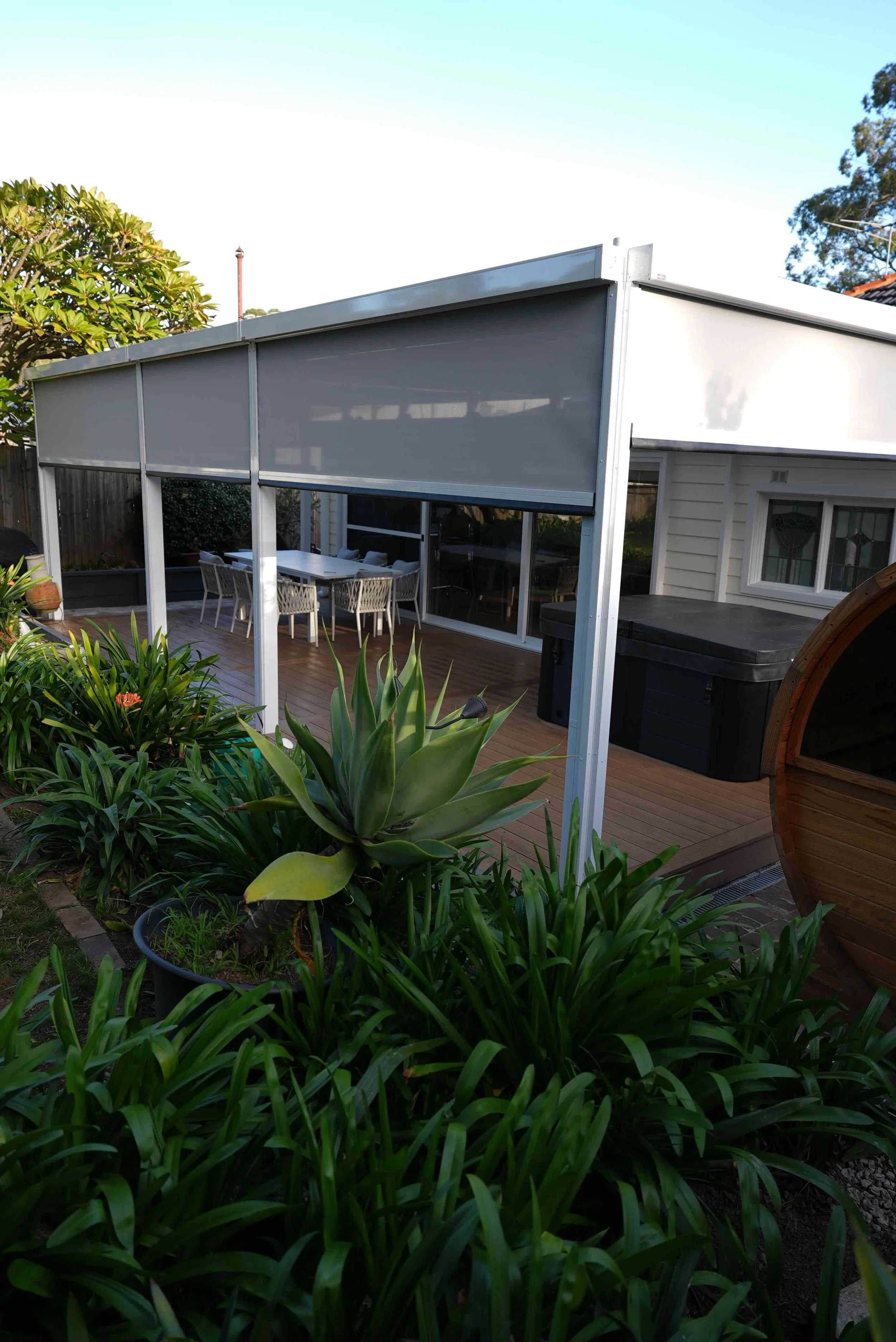 A backyard deck with a covered patio area, surrounded by lush green plants and trees, and a clear blue sky overhead.