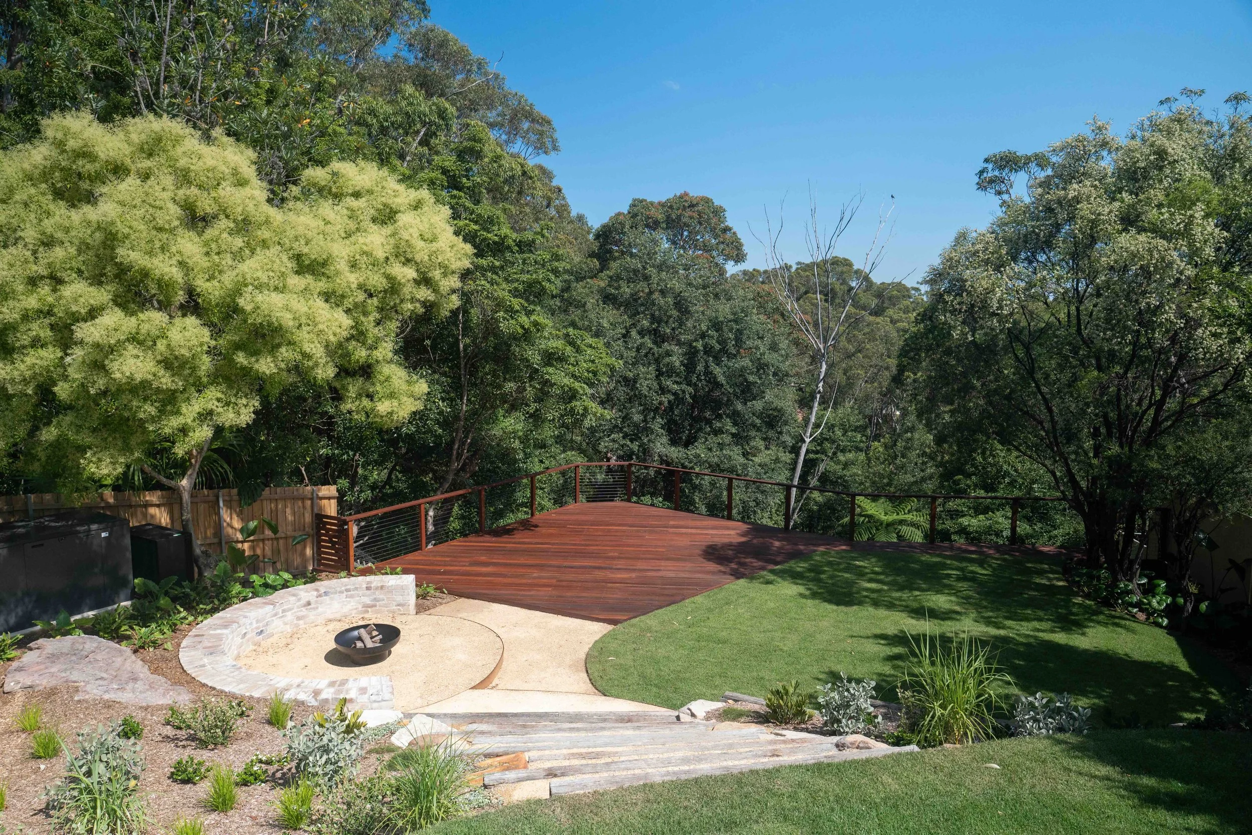 A backyard with a grassy area, a fire pit, a paved walkway, a wooden deck with a railing, lush green trees, and clear blue sky.