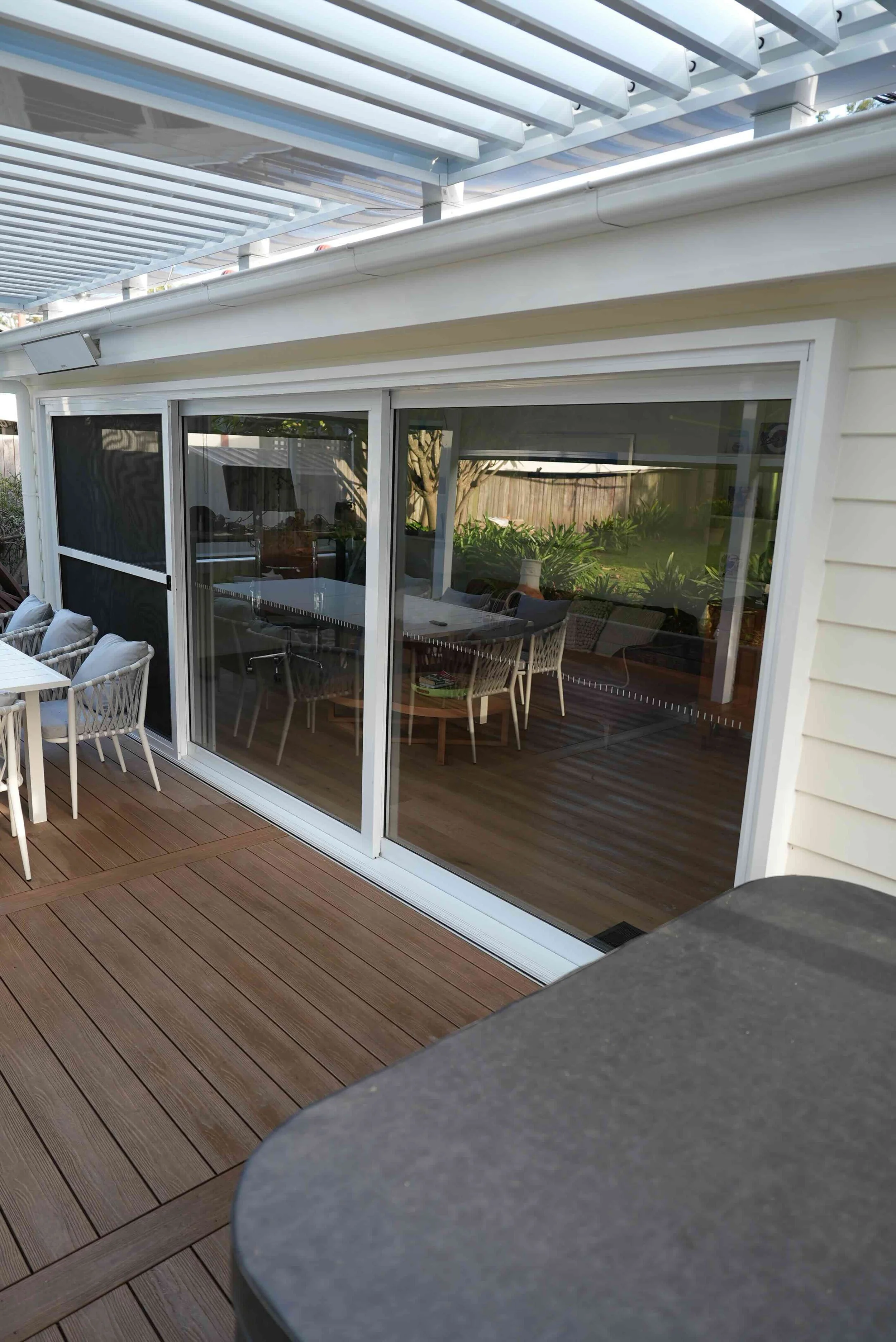 An outdoor patio with wooden decking, white chairs, and a sliding glass door leading into a room with a dining table and chairs, surrounded by a backyard with trees and plants.