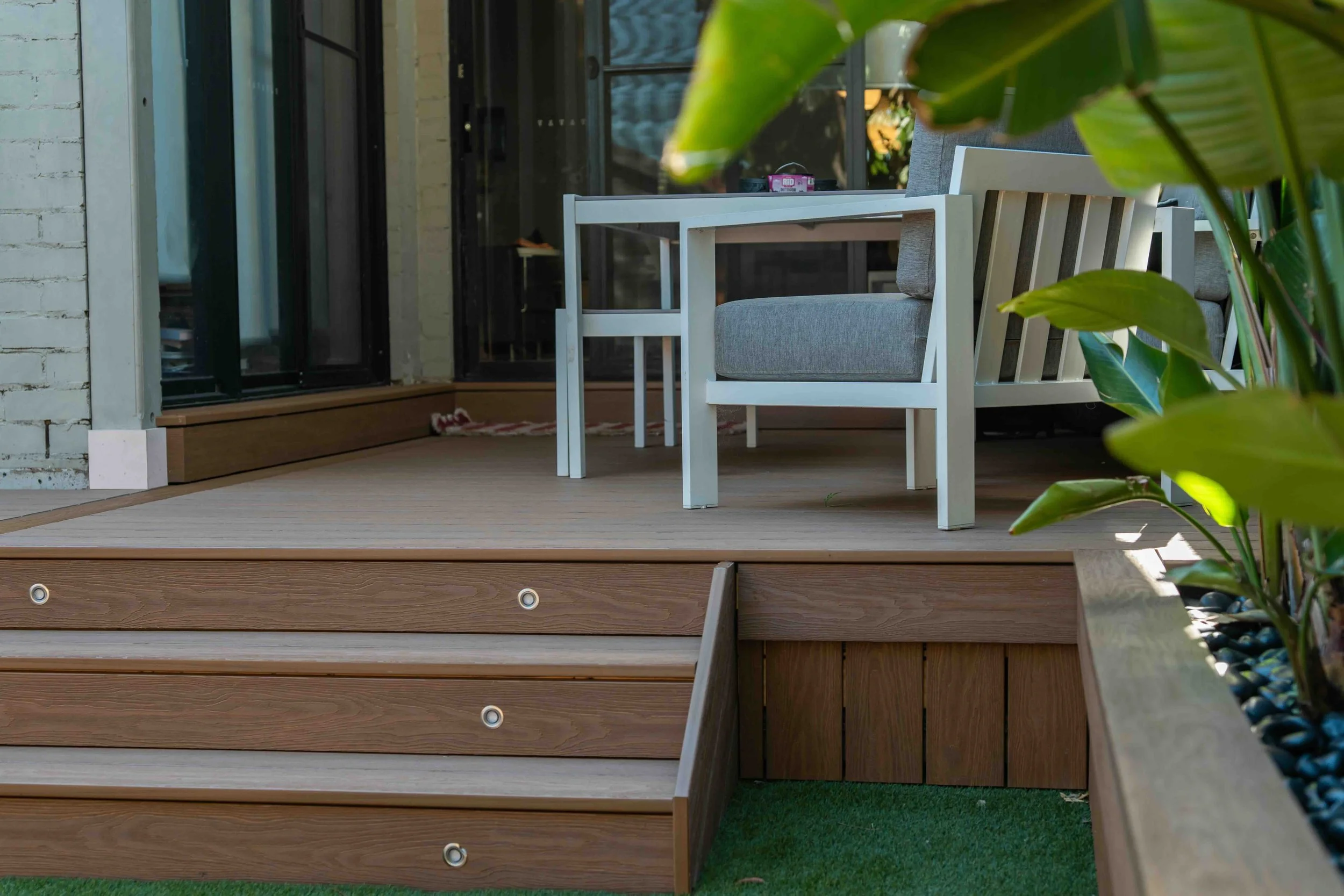 A wooden deck with a white outdoor chair and table set, partly obscured by green leaves in the foreground, and a sliding glass door leading inside a house.