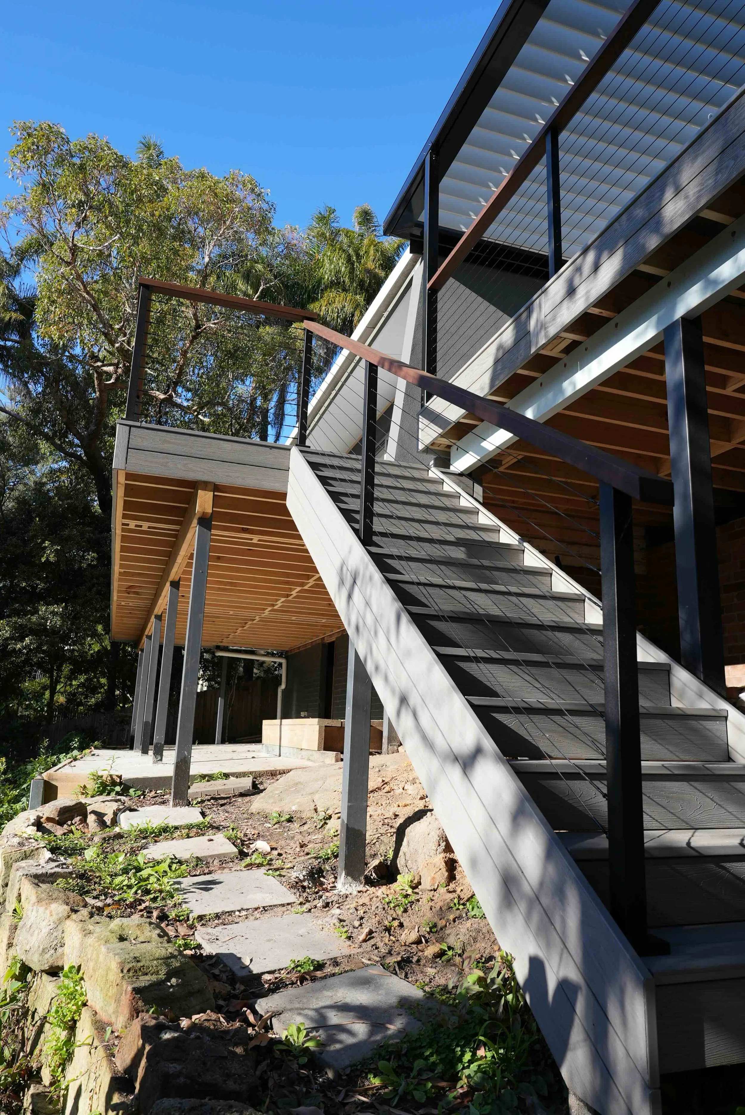 The image shows an outdoor staircase with modern design, featuring black metal railings and wooden steps. The staircase is attached to a building under construction, with parts of the structure exposed. Surrounding the area are green trees and a blue