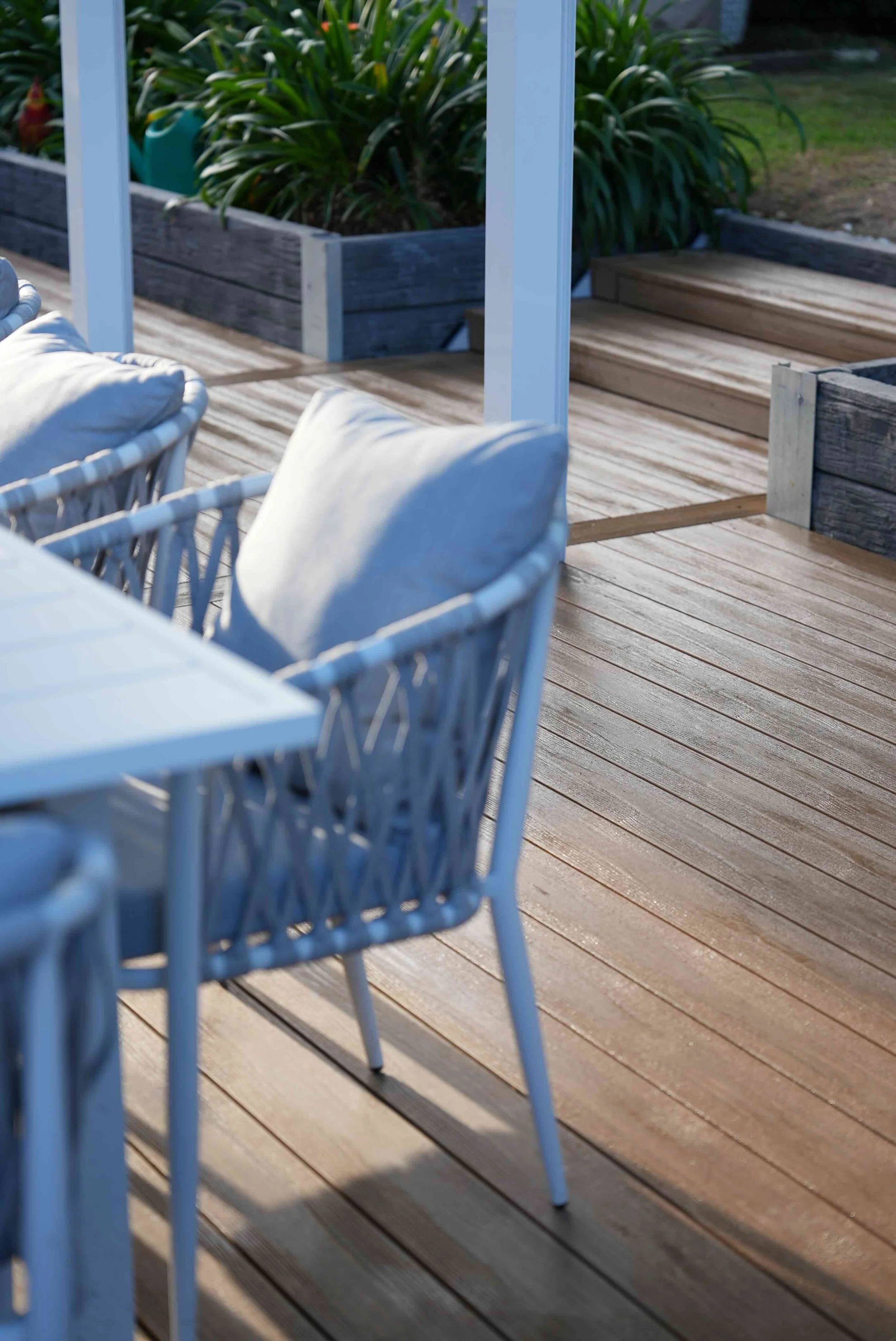 A wooden deck with outdoor furniture, including a wicker chair with white cushions, and planters with green plants in the background.