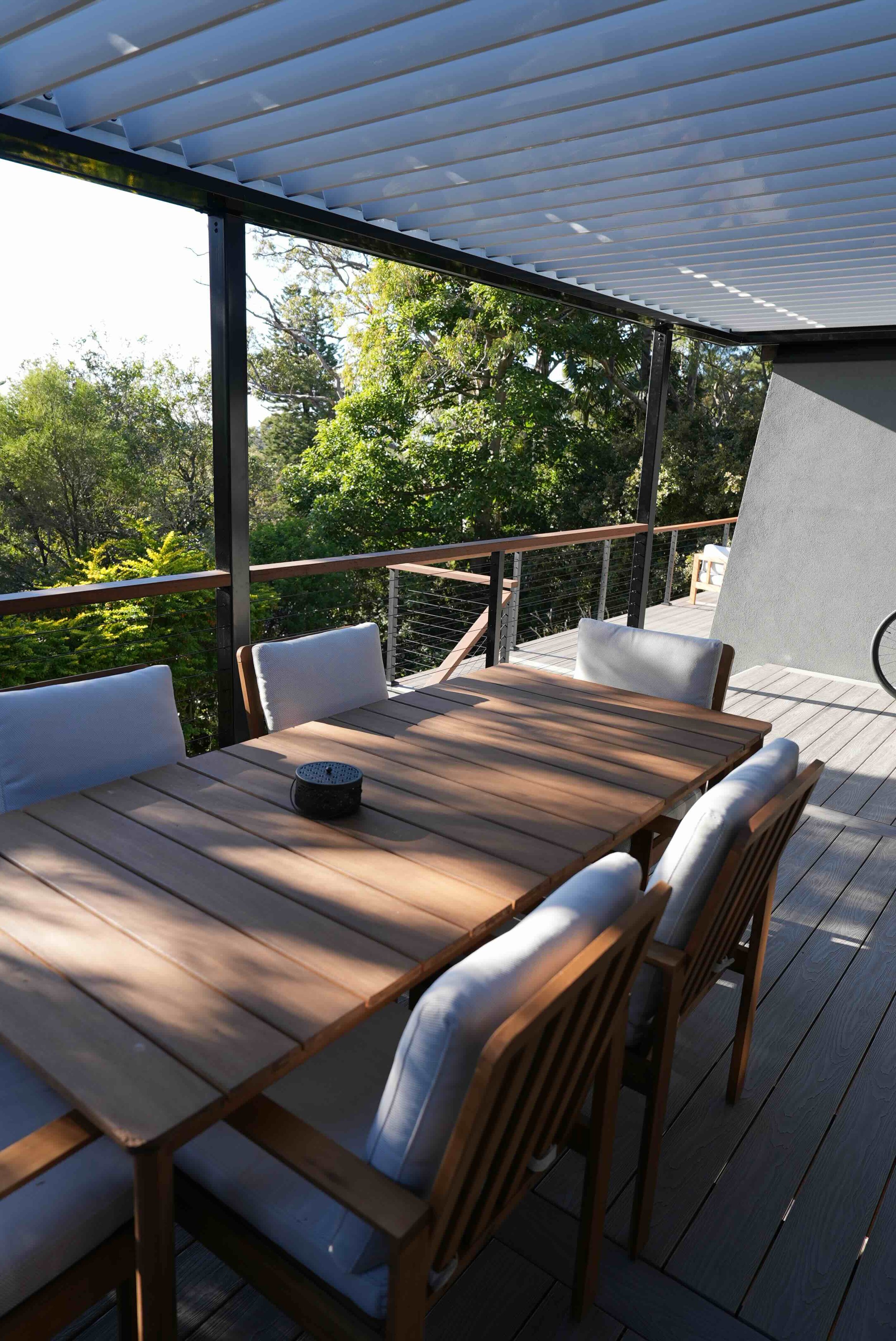 Outdoor dining area on a balcony with a wooden table and white chairs, surrounded by a railing, under a slatted roof, with lush green trees in the background.