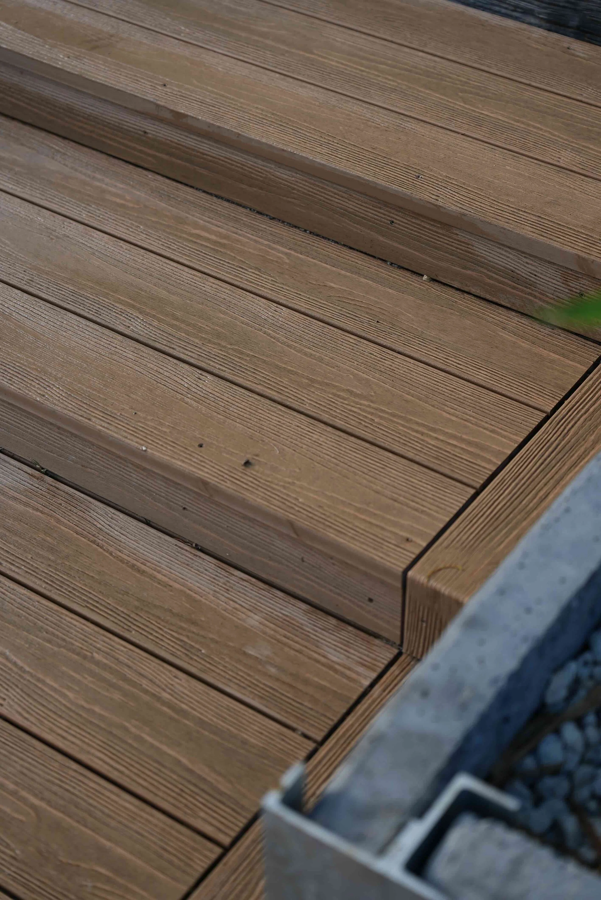 Close-up view of a wooden deck with horizontal planks, showing the wood grain texture and screws, next to a concrete surface with a metal railing.