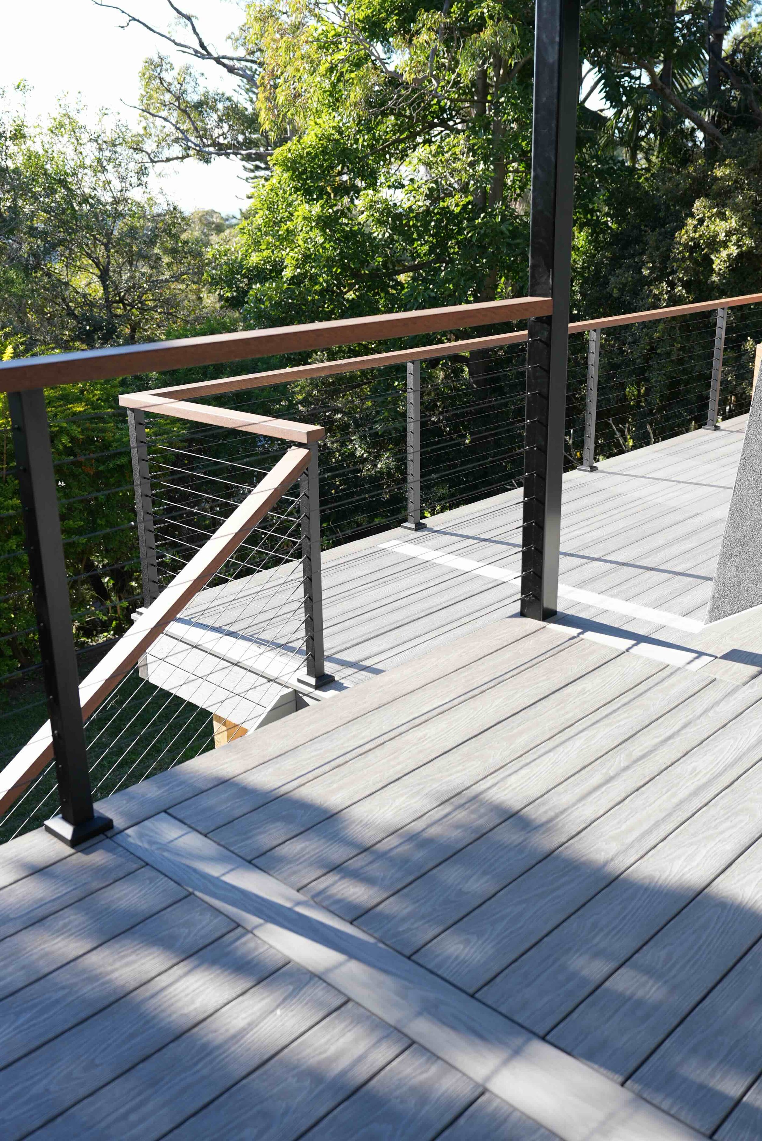 A modern wood and metal balcony with a metal wire railing and a wooden handrail, overlooking a lush green tree-filled landscape on a sunny day.