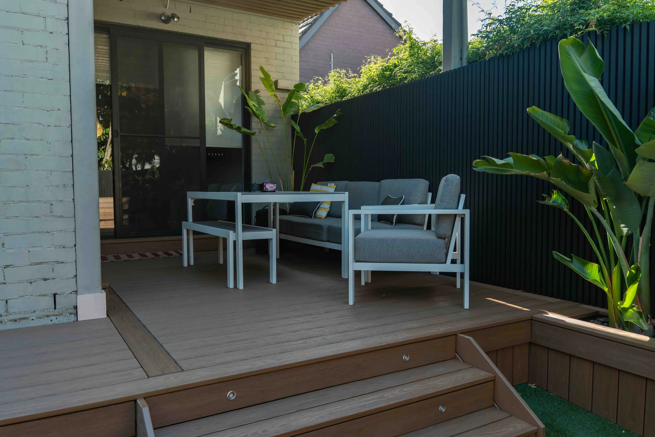 A cozy outdoor patio with a white table, gray cushioned seating, and plants against a wooden and brick wall under a slanted roof.