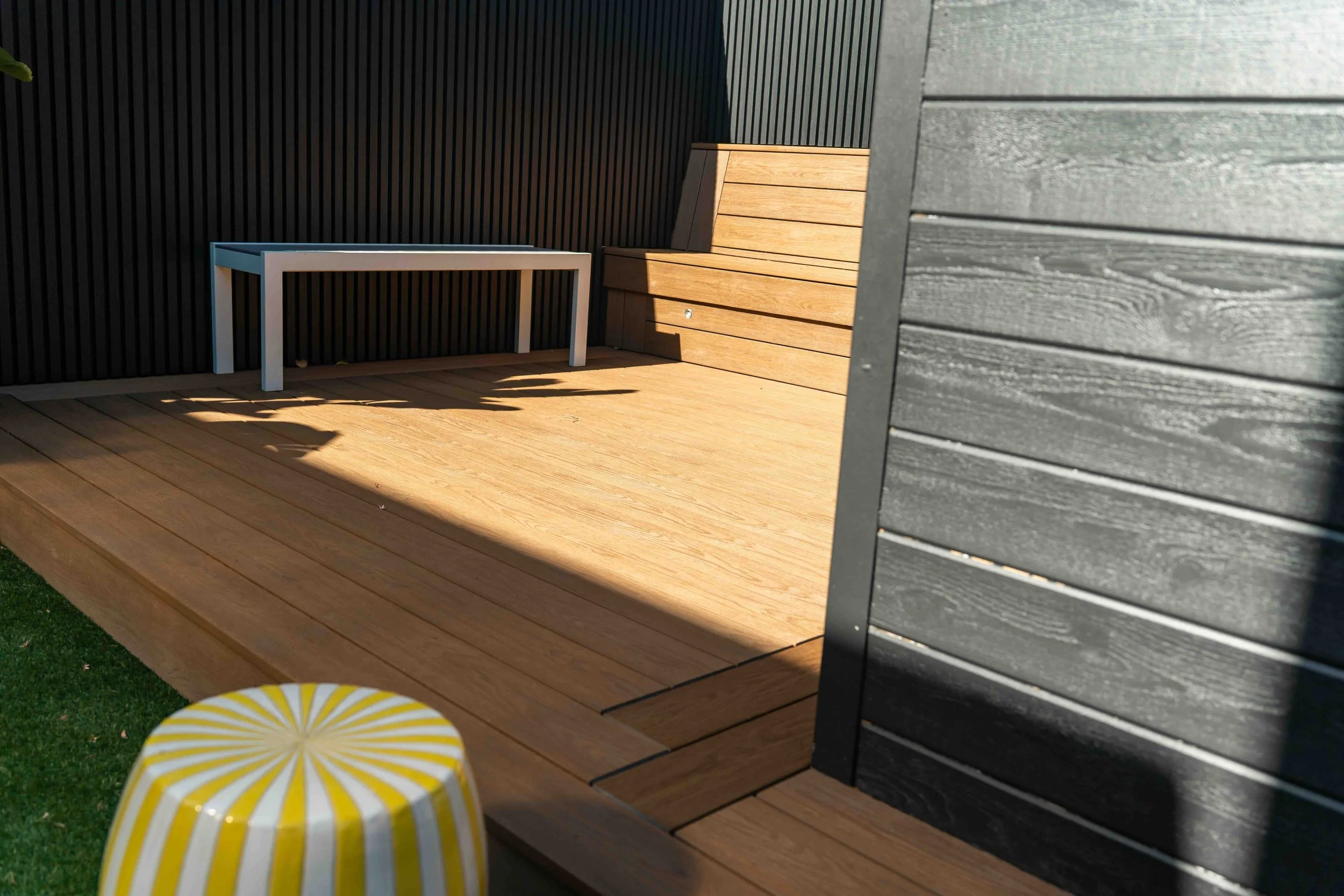 Outdoor wooden deck area with a wooden bench, a black vertical slat wall, and a dark gray wall. A yellow and white patterned stool is visible in the lower left corner.