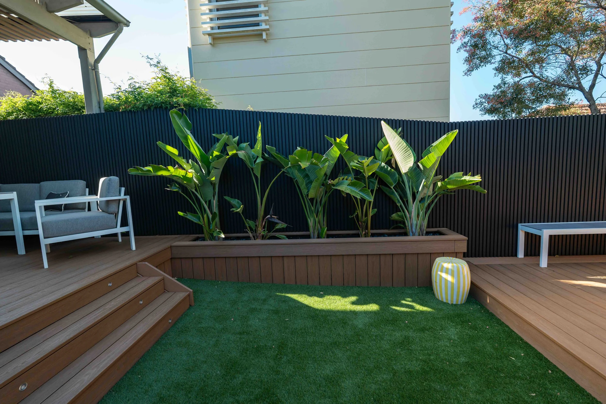 A backyard patio with wooden decking, green grass, a variety of plants, outdoor furniture including a gray couch and a white chair, a black fence, and trees in the background.