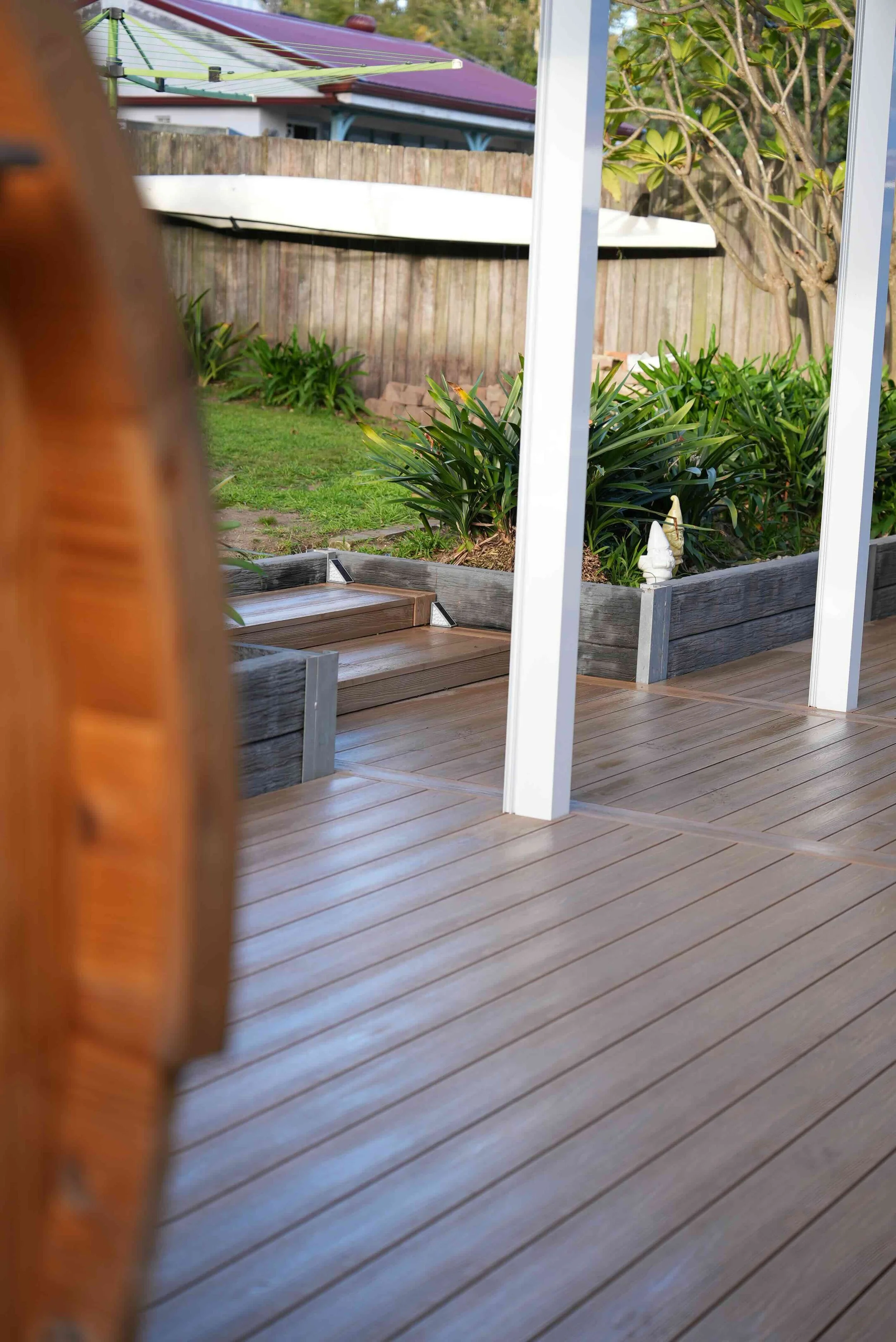 View of a backyard with a wooden deck, garden plants, and a white patio structure. Part of a house with a red roof and a boat above the fence are visible in the background.