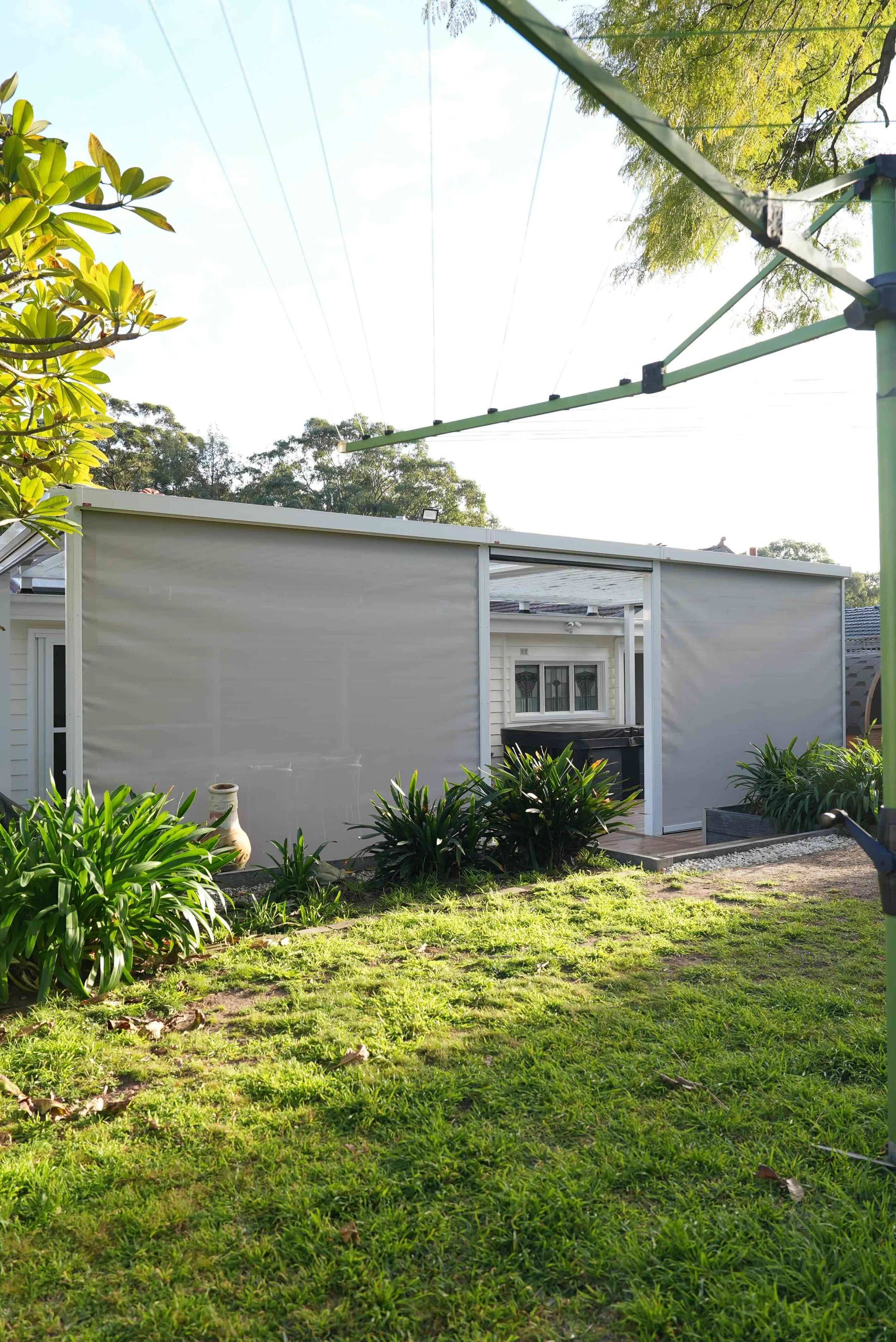 A backyard scene with green grass, bushes, and trees. There is a small wooden deck with a white building and a sliding door or window. A green rotary clothesline is stretching across the sky, which is bright and partly cloudy. The yard has lush, heal