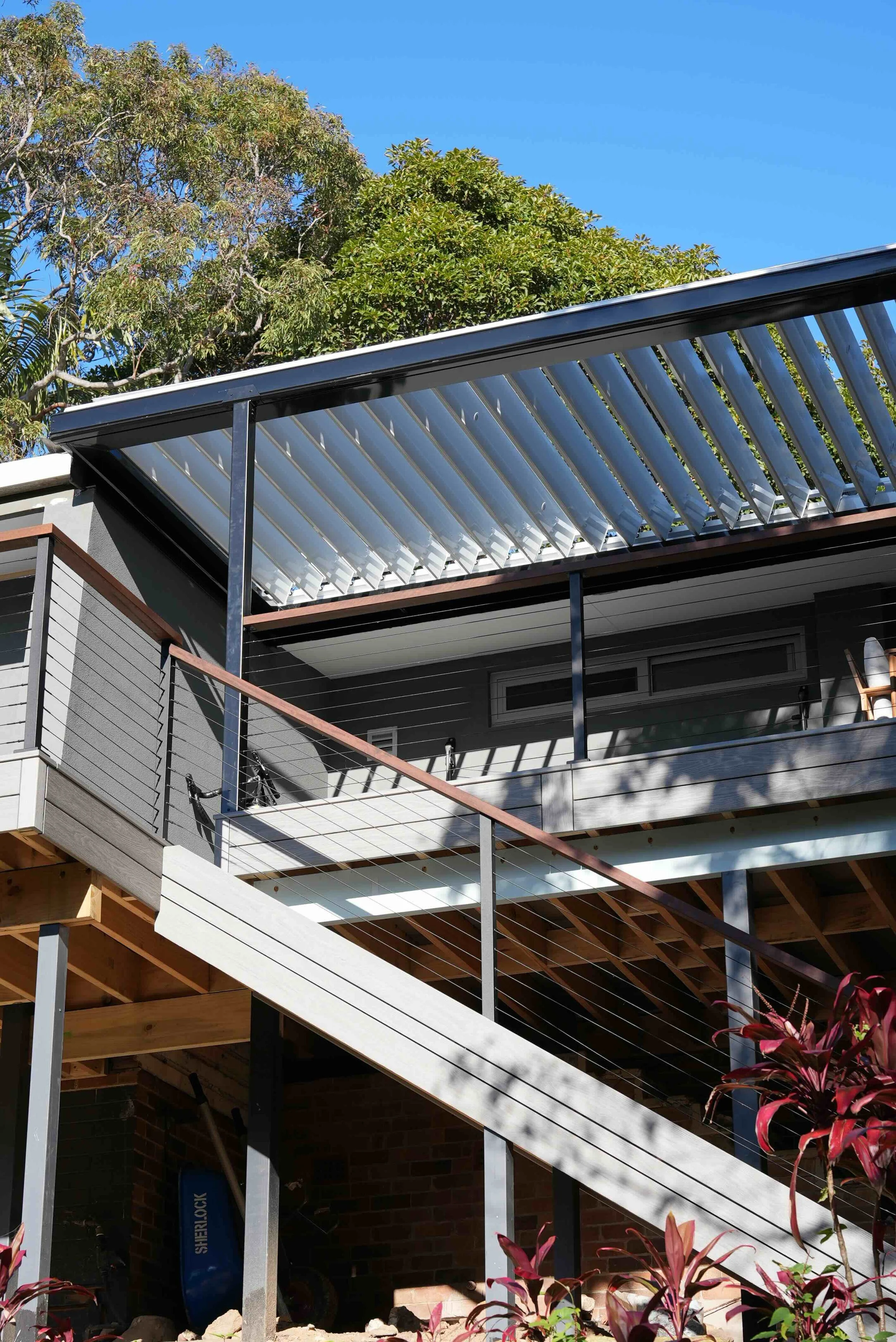 A multi-story building under construction with exposed wooden and metal framework, a metal balcony, and a solar panel installed on the roof, with trees and a clear blue sky in the background.