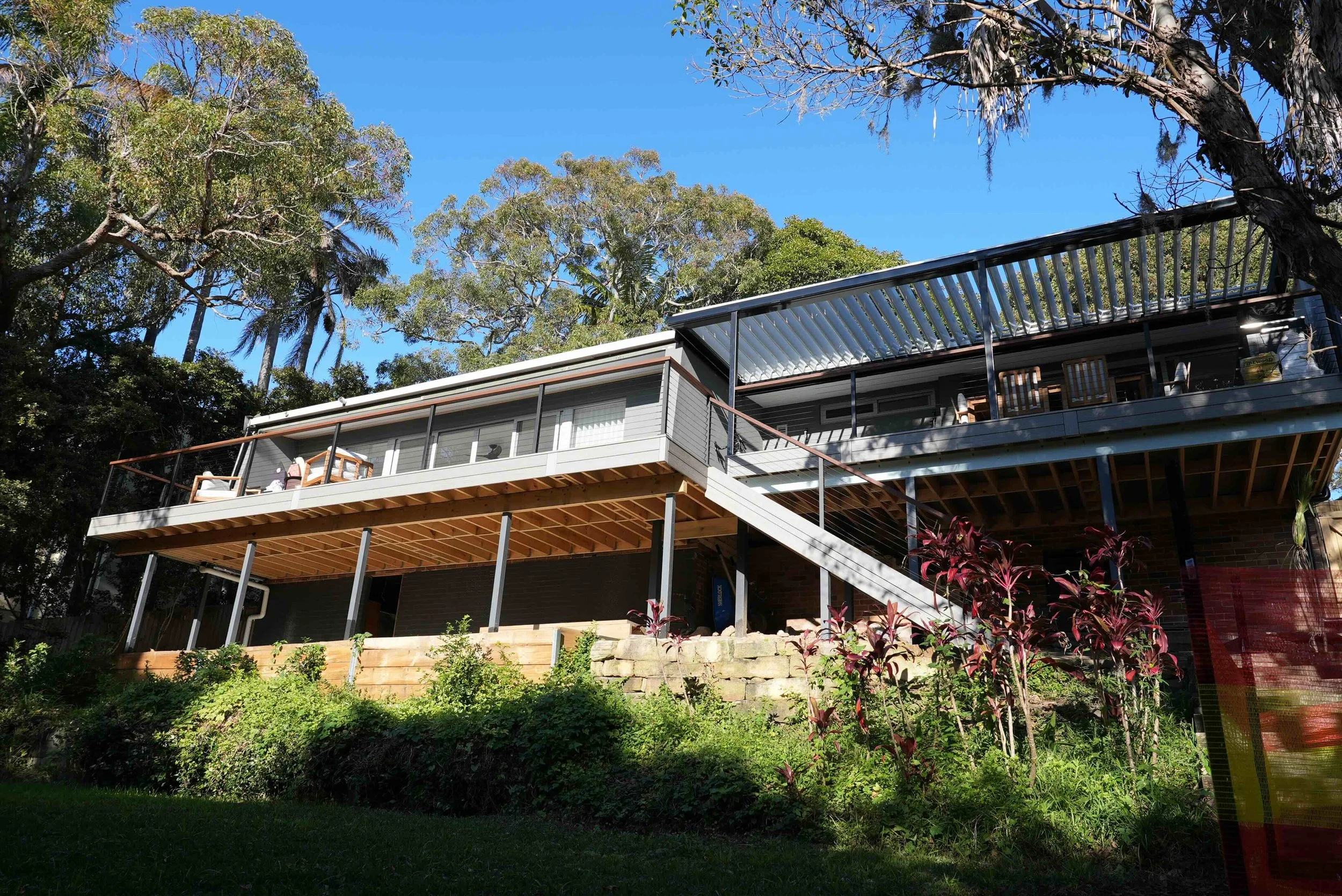 A modern house on stilts with a large balcony surrounded by trees and plants, under a clear blue sky.