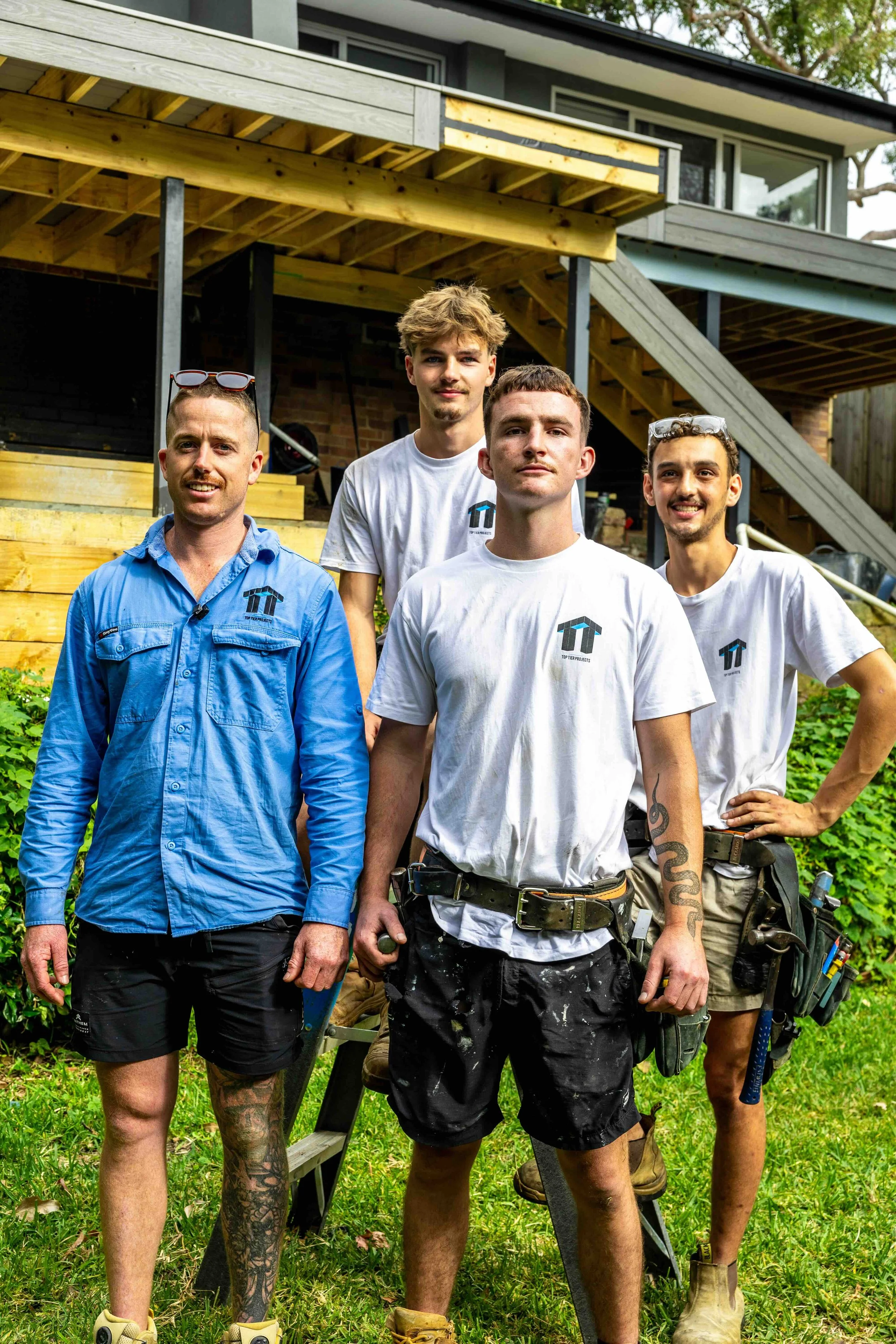 Four men standing outdoors in front of a house under construction, wearing casual work clothes and safety glasses, with tools hanging from their belts.
