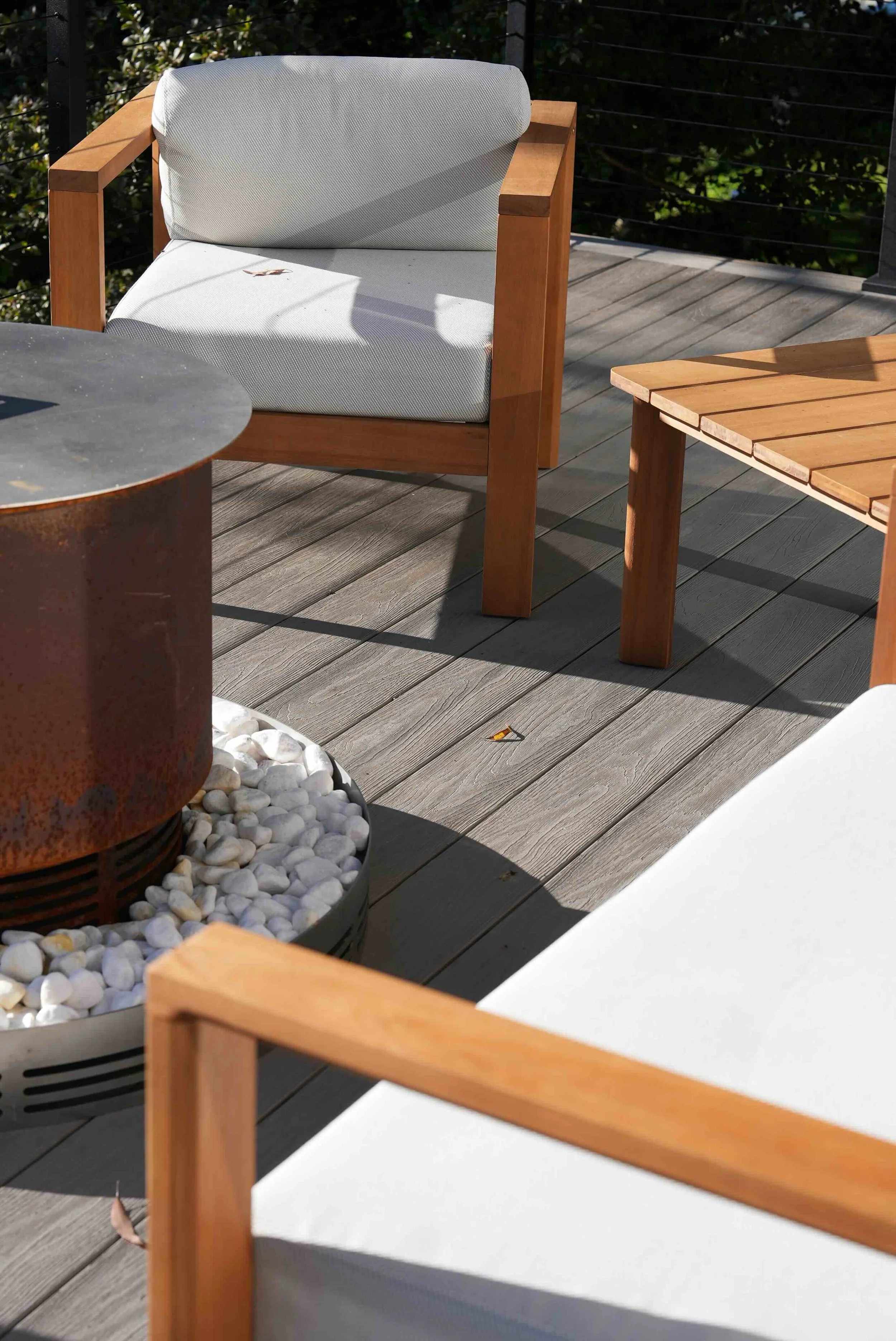 Outdoor patio area featuring a wooden chair with a white cushion, a wooden table, a fire pit filled with white stones, and a second wooden chair. Shaded by sunlight, surrounded by tree foliage and a black railing.