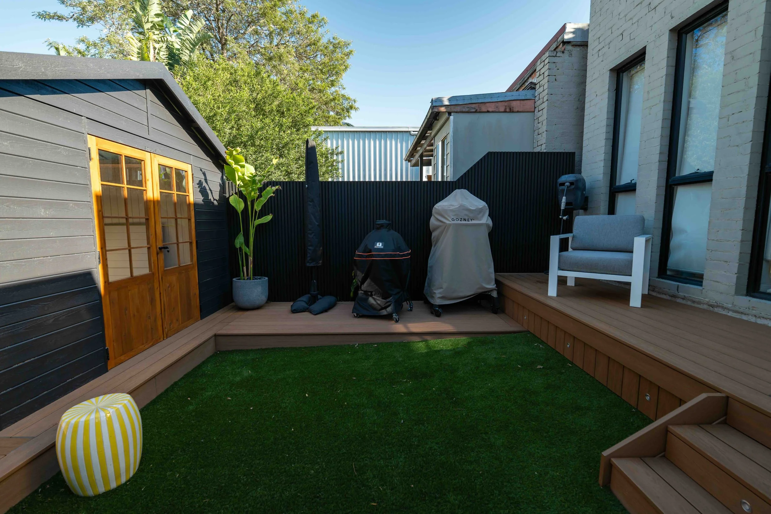 A backyard patio with a wooden deck, a small green lawn, a black picket fence, a gray outdoor chair, two covered barbecue grills, a large potted plant, and a yellow-striped outdoor cushion. There are trees and a building with windows in the backgroun