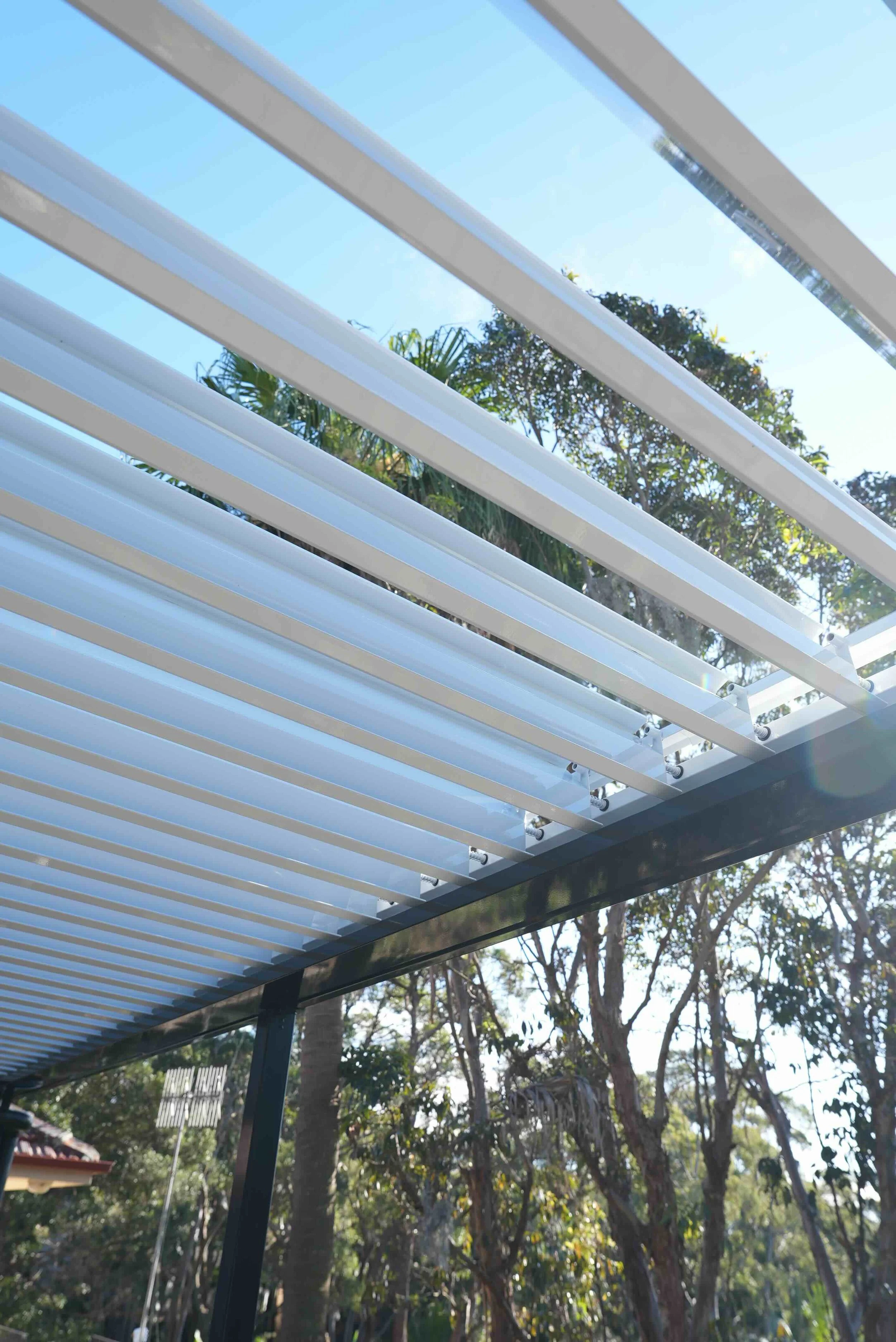Close-up of white horizontal slats on a modern outdoor pergola with tree branches and blue sky visible in the background.