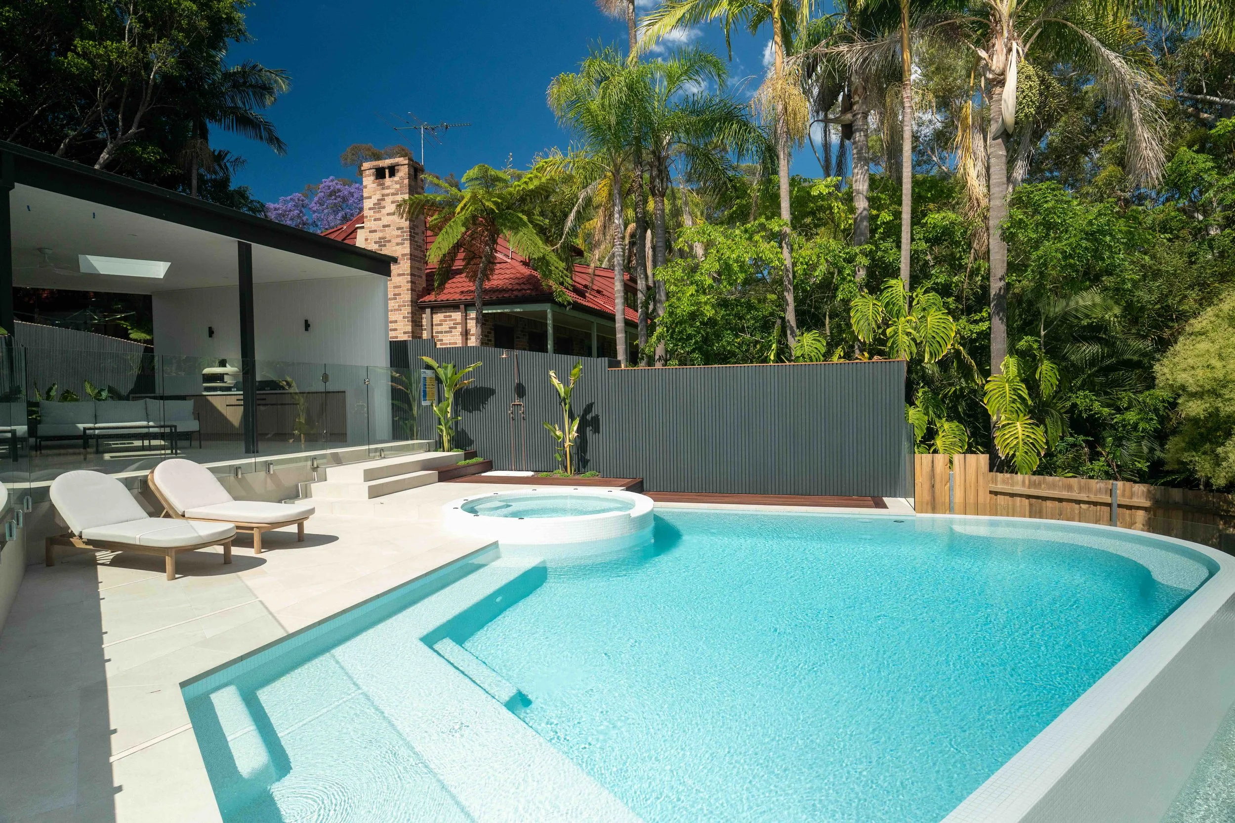 Modern backyard with a kidney-shaped swimming pool, lounge chairs, lush green trees, and a house with a red roof and chimney.