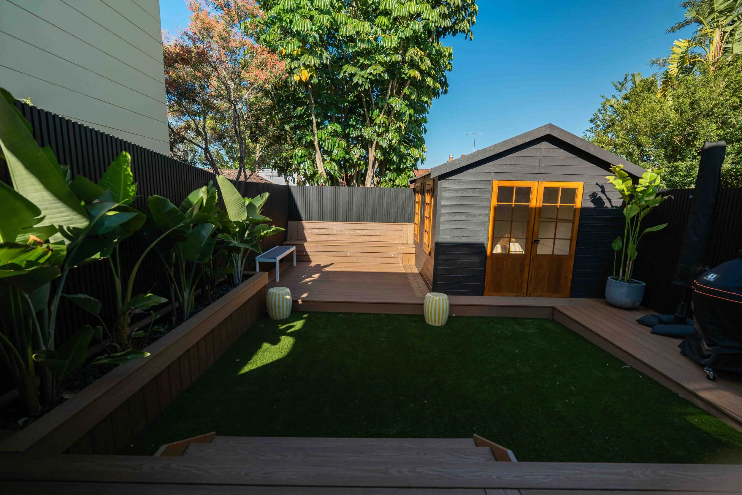 A backyard with a wooden deck, green grass, plants along the fence, a small black and wood shed with windows, and tall trees with green and pink leaves under clear blue sky.