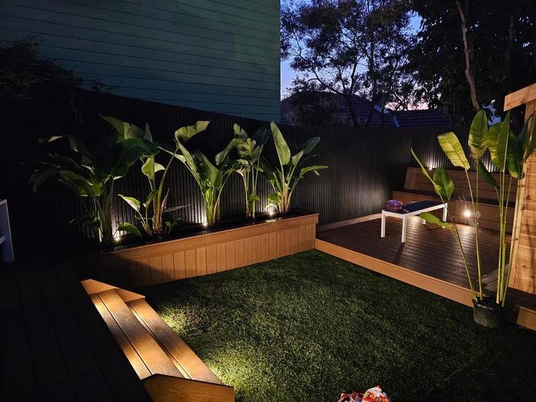 Night view of a backyard patio with a wooden deck, potted tropical plants, a small white table, and outdoor lighting, with trees in the background.