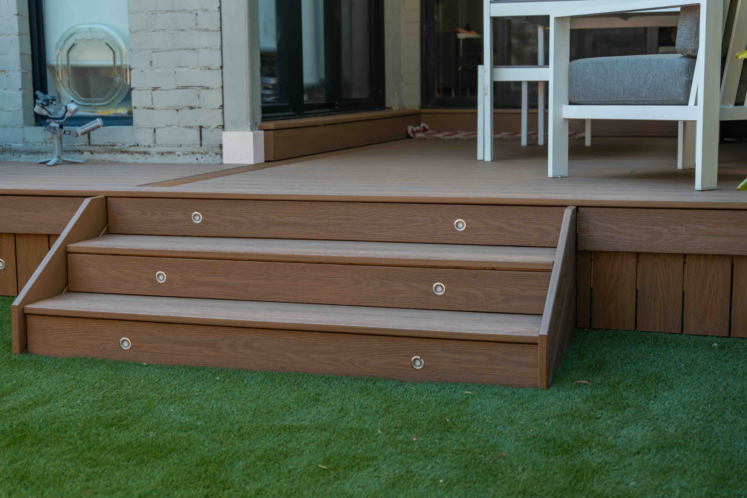 A backyard scene featuring a raised wooden deck with a set of stairs and a grassy lawn. There is a white chair with gray cushions on the deck and a white frame structure partially visible. The background shows a brick wall and glass doors.